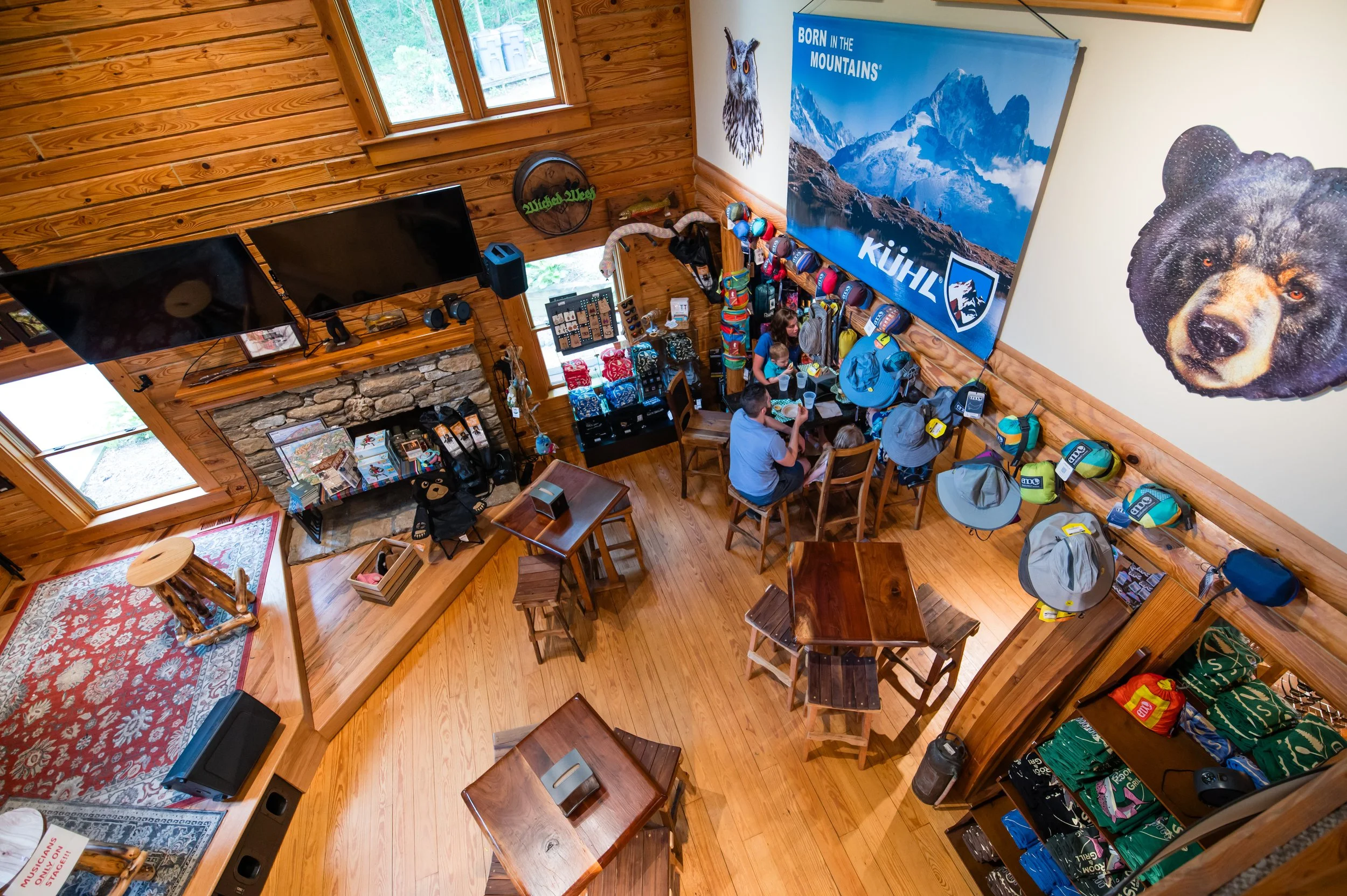 Interior of a wooden lodge decorated with outdoor and mountain-themed items, including a large mountain poster, shelves with backpacks and gear, a stone fireplace, and people sitting at a table.