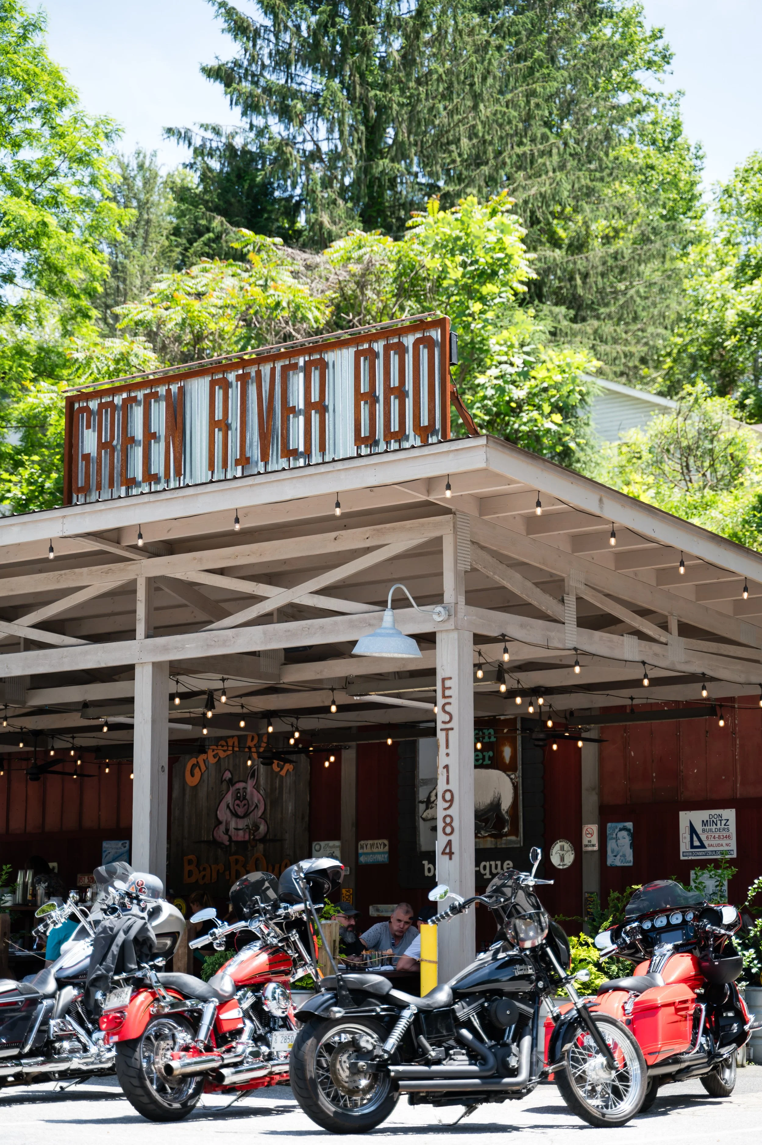 Exterior view of Green River BBQ, a barbecue restaurant with an outdoor seating area. There are multiple motorcycles parked in front, and people are sitting and enjoying their meals under a covered patio. The background features lush green trees and a clear blue sky.