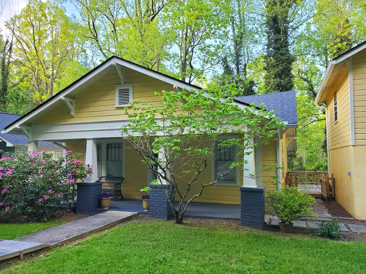 Yellow house with a porch, surrounded by green trees and plants, and a small paved walkway leading to the front door.