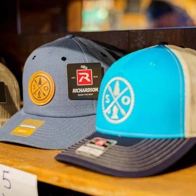 Two baseball caps on a wooden surface, one gray with a yellow patch and Richardson logo, the other blue and beige with a white logo of crossed axes and circles.