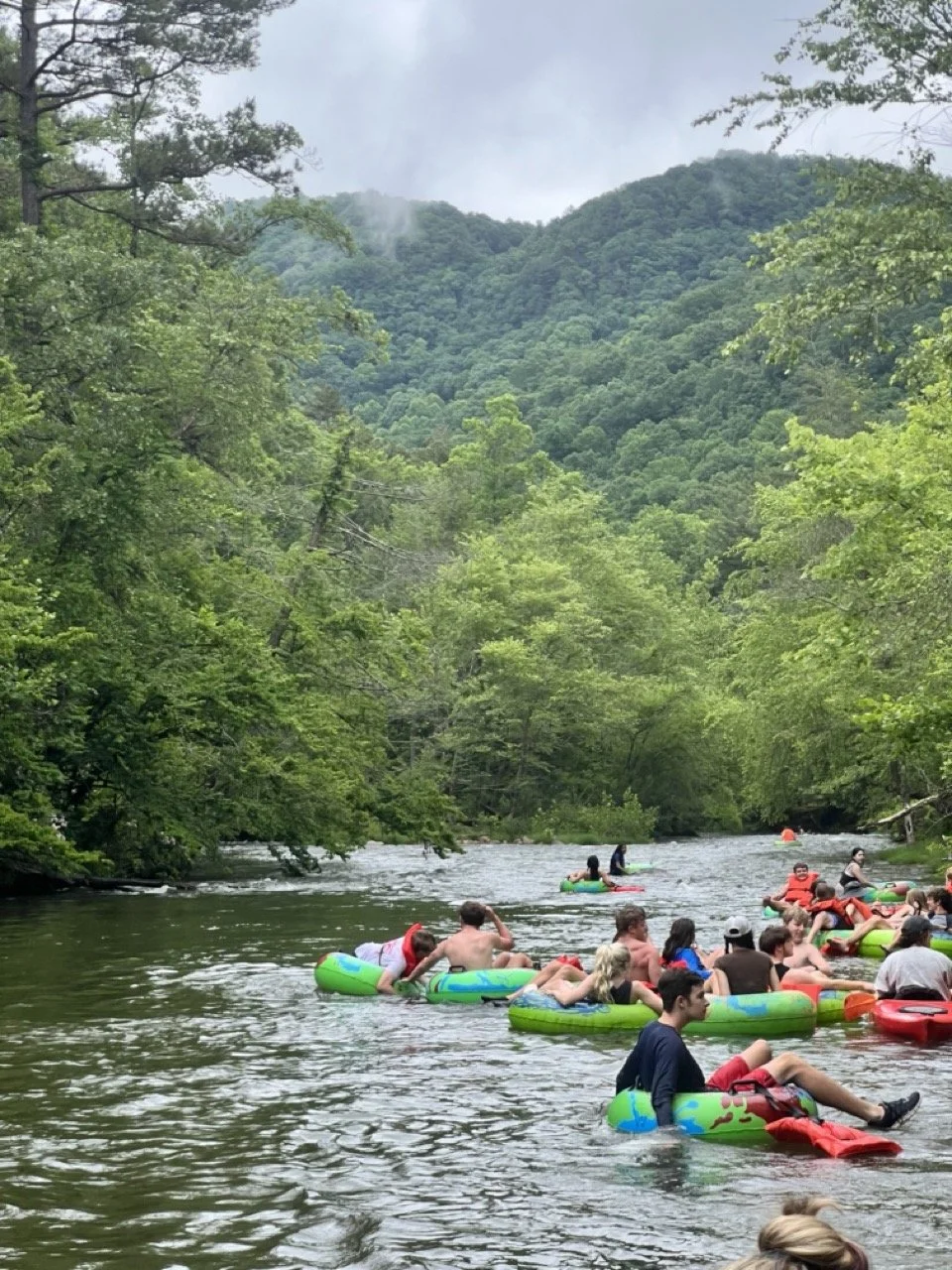 People floating on a river surrounded by lush green trees and mountains in the background.