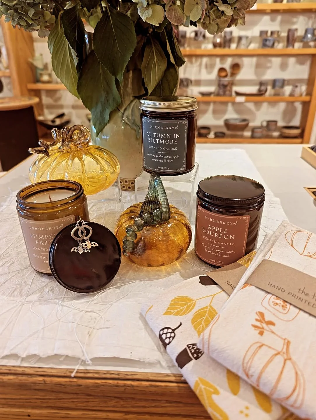 Autumn-themed decorative display with four scented candles labeled autumn in biltmore, apple bourbon, pumpkin patch, and a third candle. The display includes small decorative pumpkins, dried leaves, and a leather keychain, all arranged on a table wit