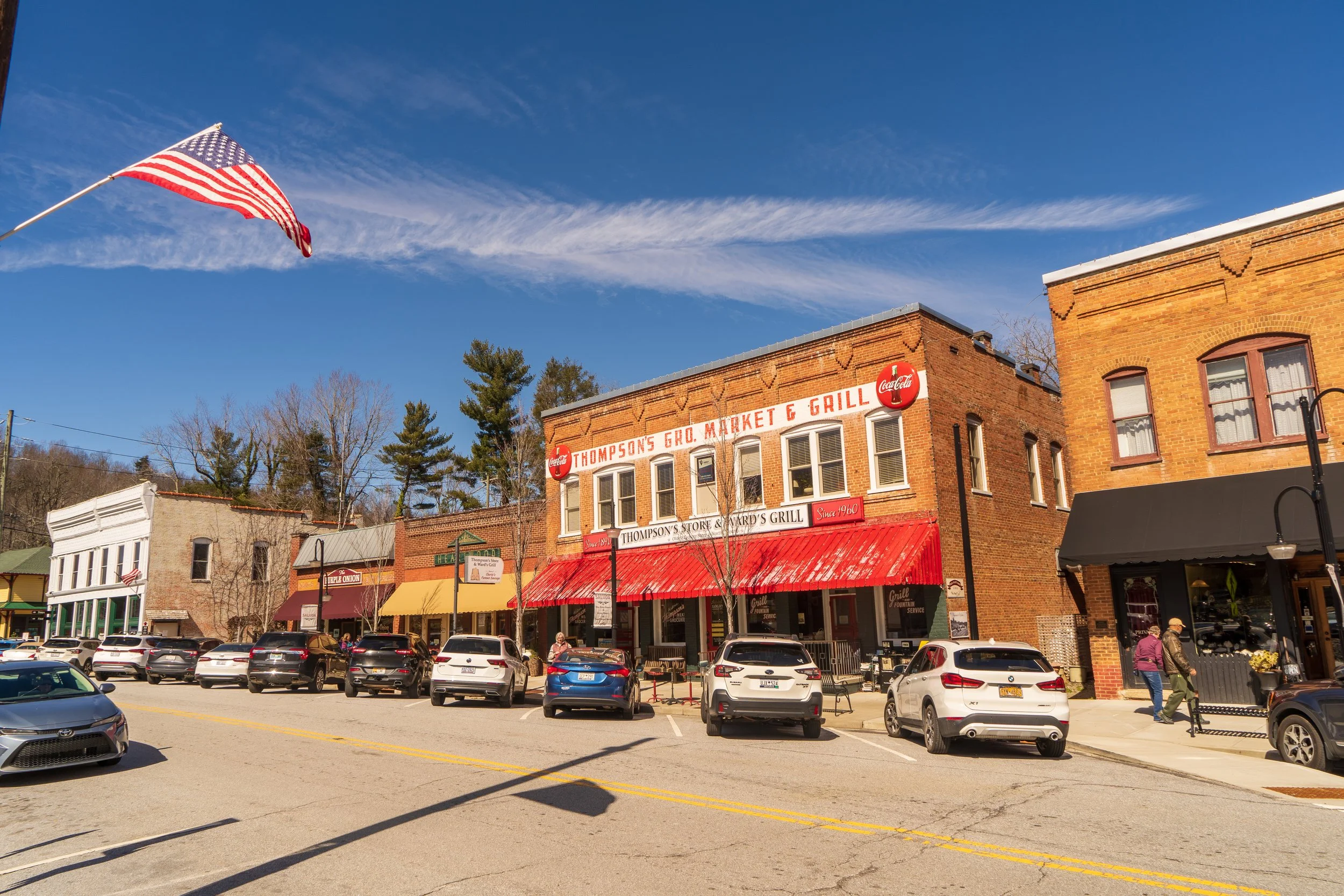 Main street in a small town with brick buildings and storefronts, parked cars, pedestrians, an American flag flying, and a partly cloudy blue sky.