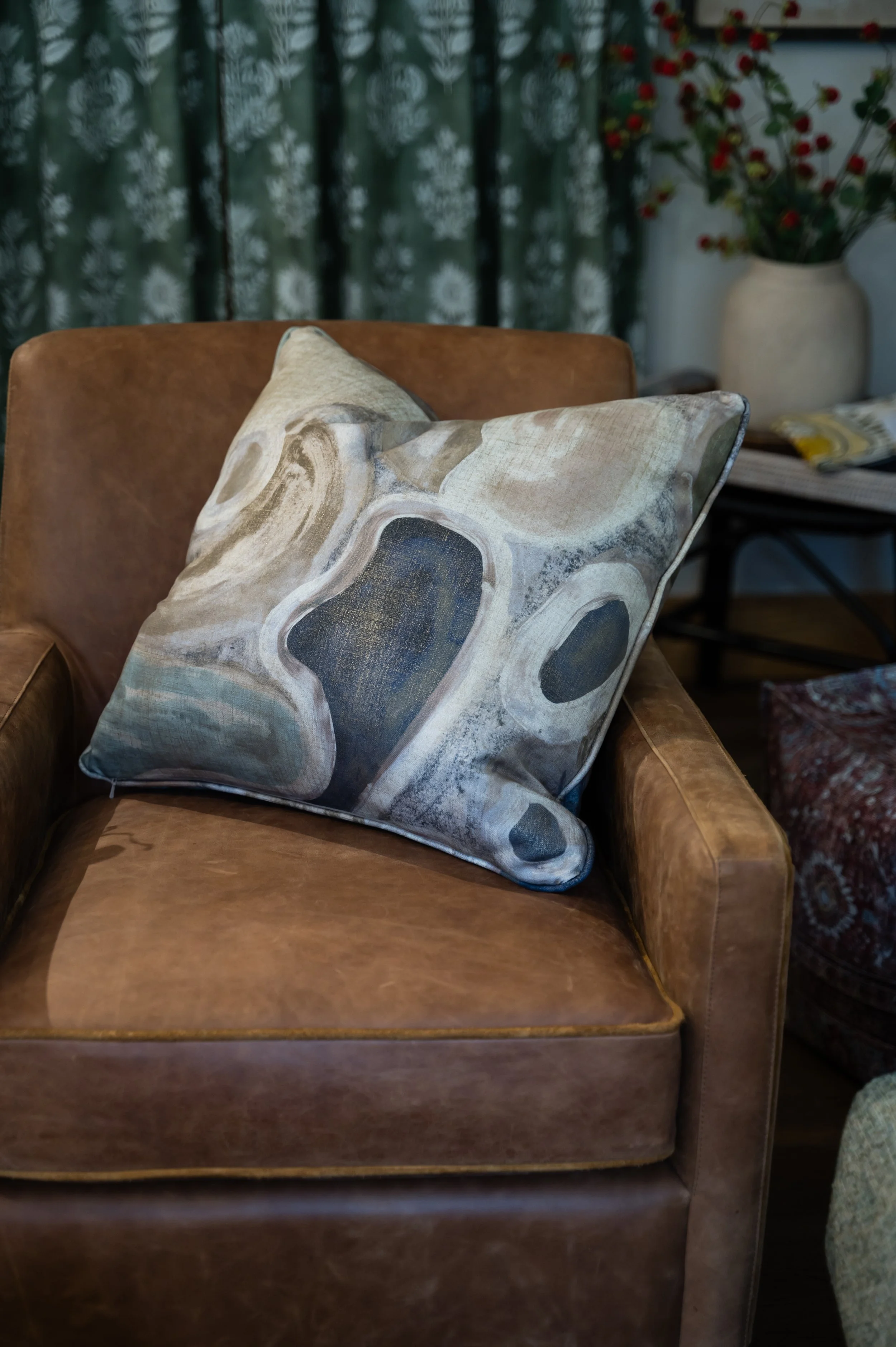 A beige and brown leather armchair with two decorative pillows, one with an abstract pattern, situated in a cozy room with dark floral curtains and a large vase of red berries in the background.