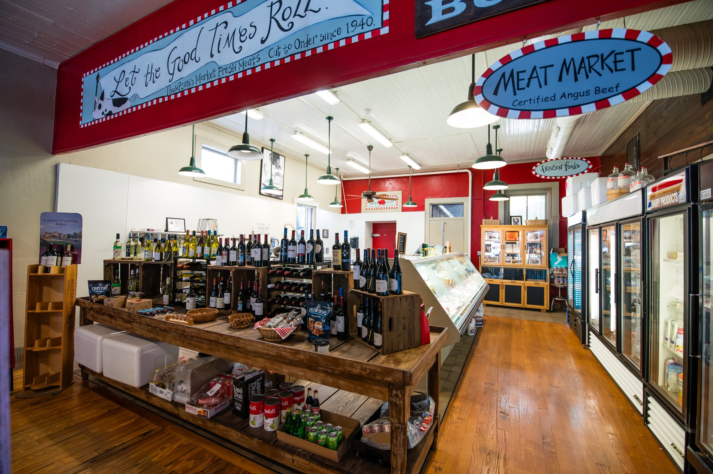 Interior view of a store with shelves of bottled wine, canned drinks, and other grocery items, featuring signs for a meat market and frozen foods. The ceiling has green hanging lights, and the floor is wooden.