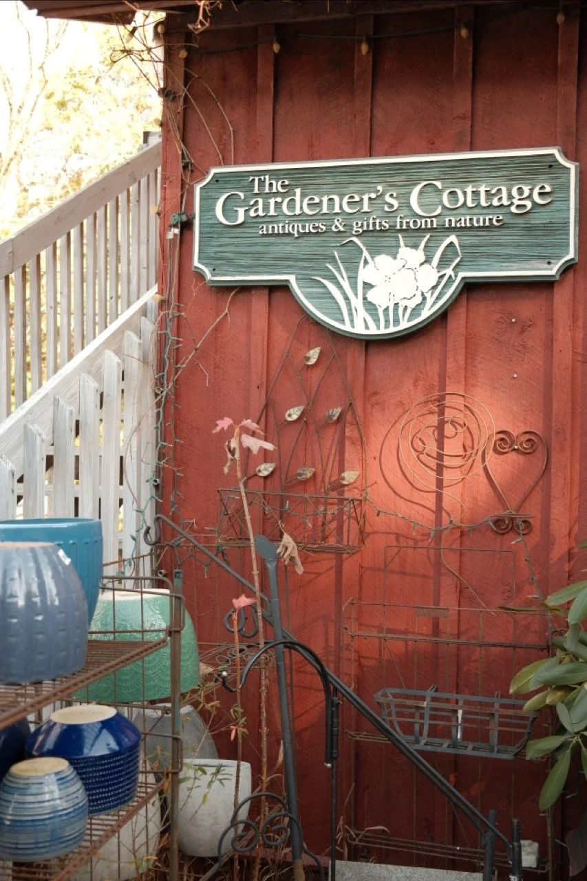 Sign for The Gardener's Cottage shop with antiques and gifts from nature, mounted on a red wooden wall outside a shop, surrounded by metal garden ornaments and pottery
