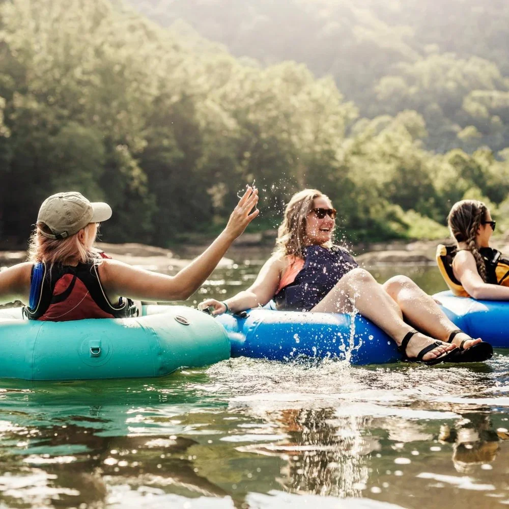 Three women enjoying a day on the water on inflatable tubes, with one woman splashing water and two women wearing sunglasses and life jackets.