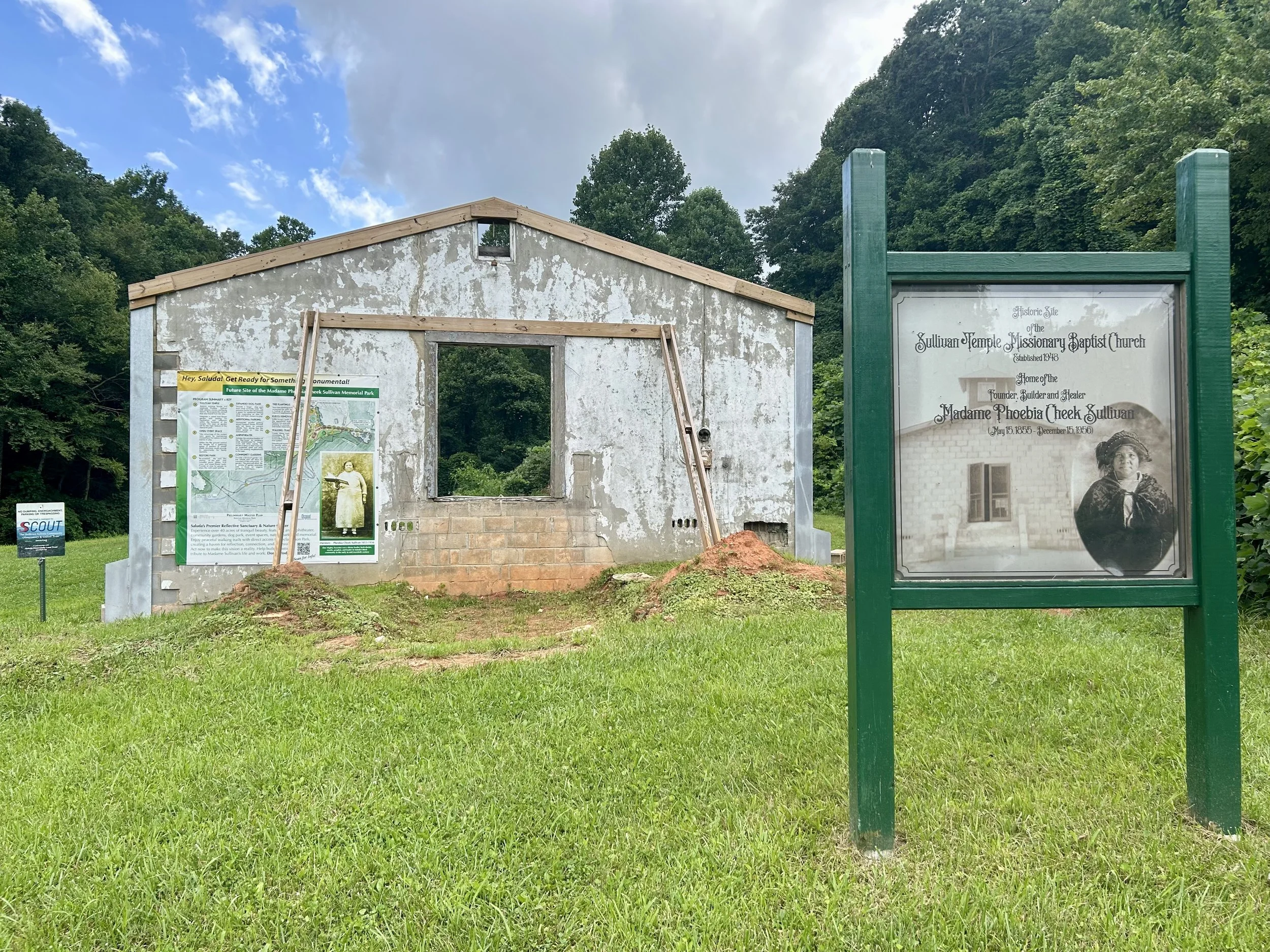 An under-construction building with unfinished walls and a window opening, surrounded by green grass and trees. There's a green-framed informational sign on the right about the Sullivan Temple Missionary Baptist Church.