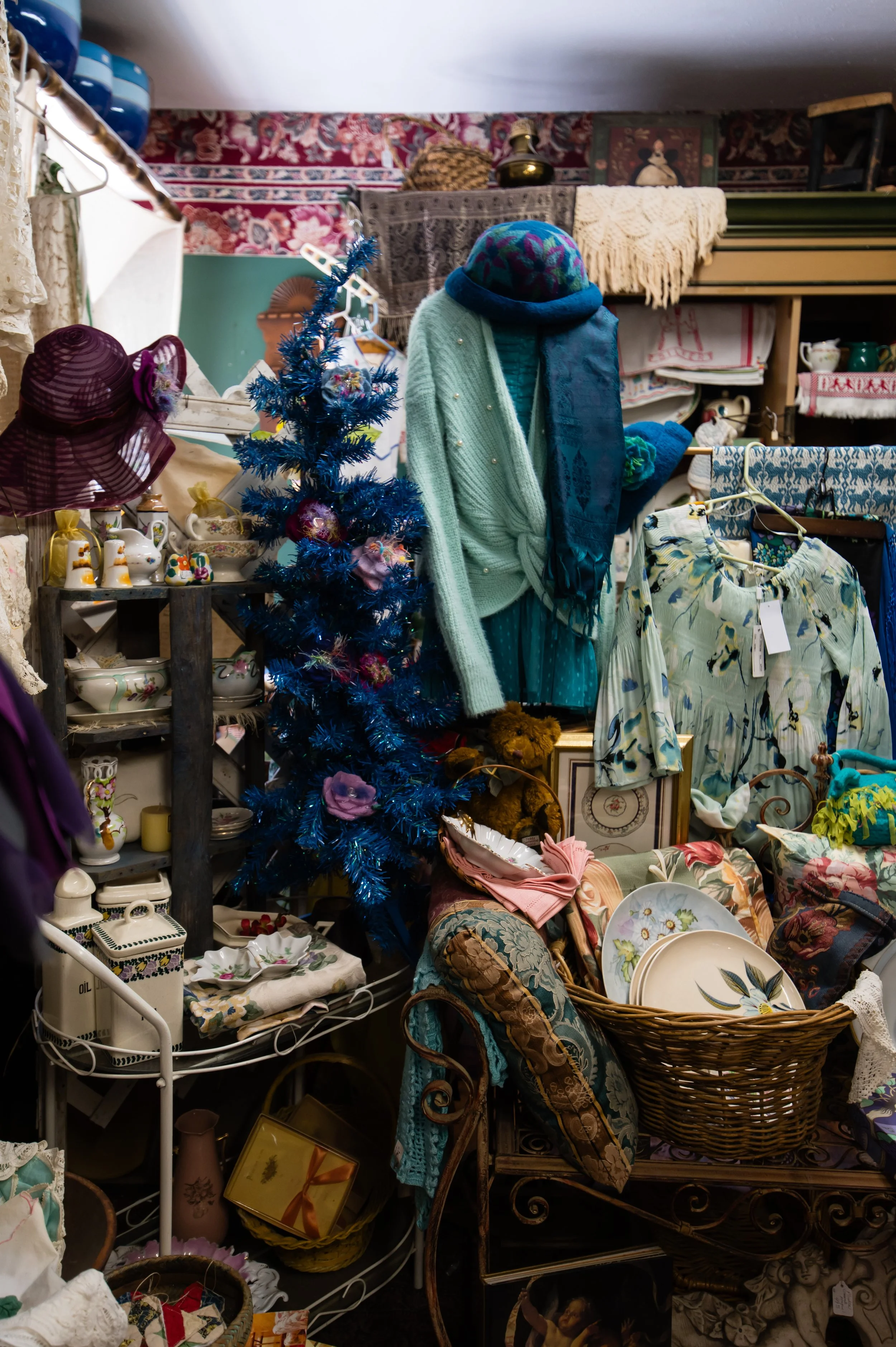 A cluttered vintage thrift store display with a small decorated blue Christmas tree, various ceramic tea sets and decorative items, clothing including a green cardigan, scarf, and dress, a wicker basket filled with fabric, and shelves with covered bo