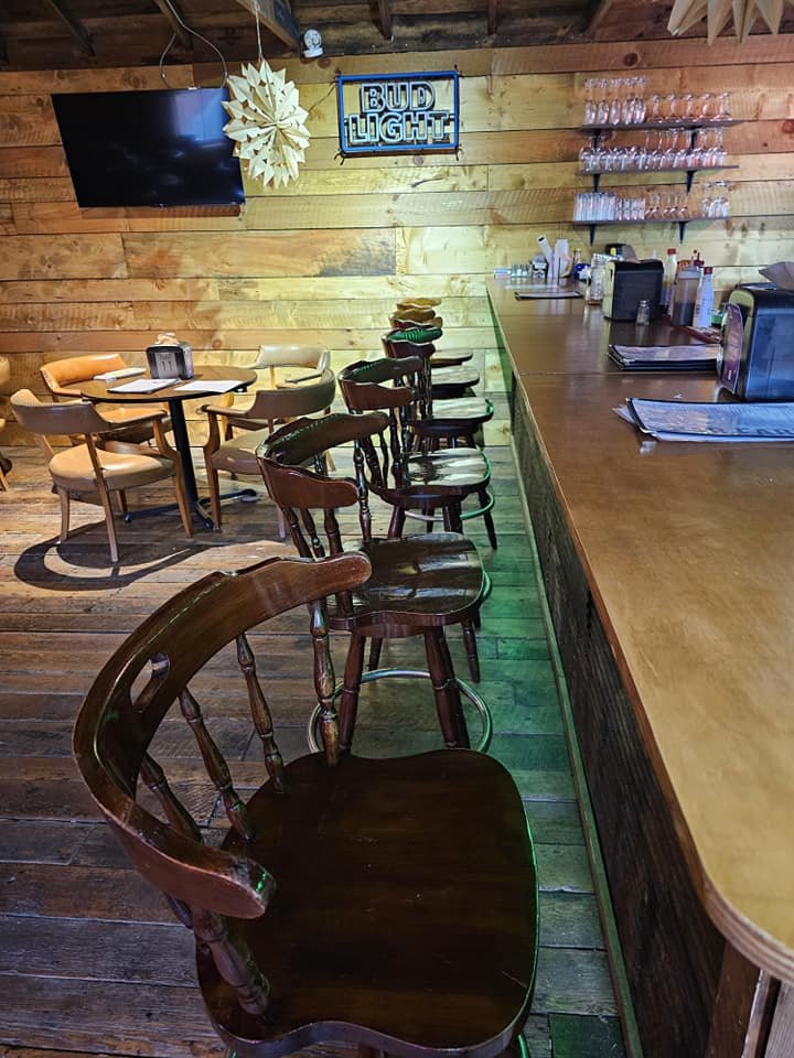 Interior of a bar or restaurant with wooden walls and floor, a row of wooden bar stools, a counter with drink glasses, and a television mounted on the wall. There is a sign that reads 'Bud Light' and decor hanging above.