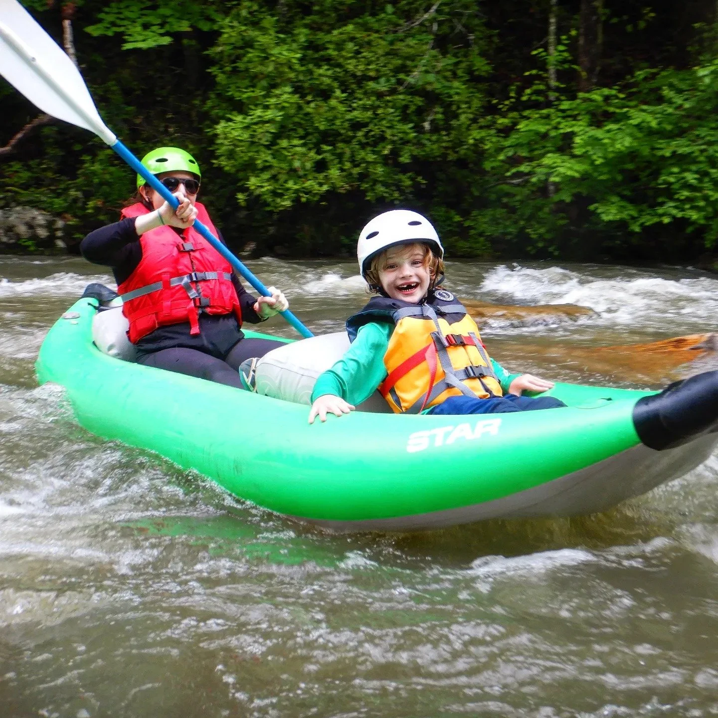 A woman and a young boy are kayaking together in a river surrounded by green trees. Both are wearing helmets and life jackets, and are paddling in a green kayak.