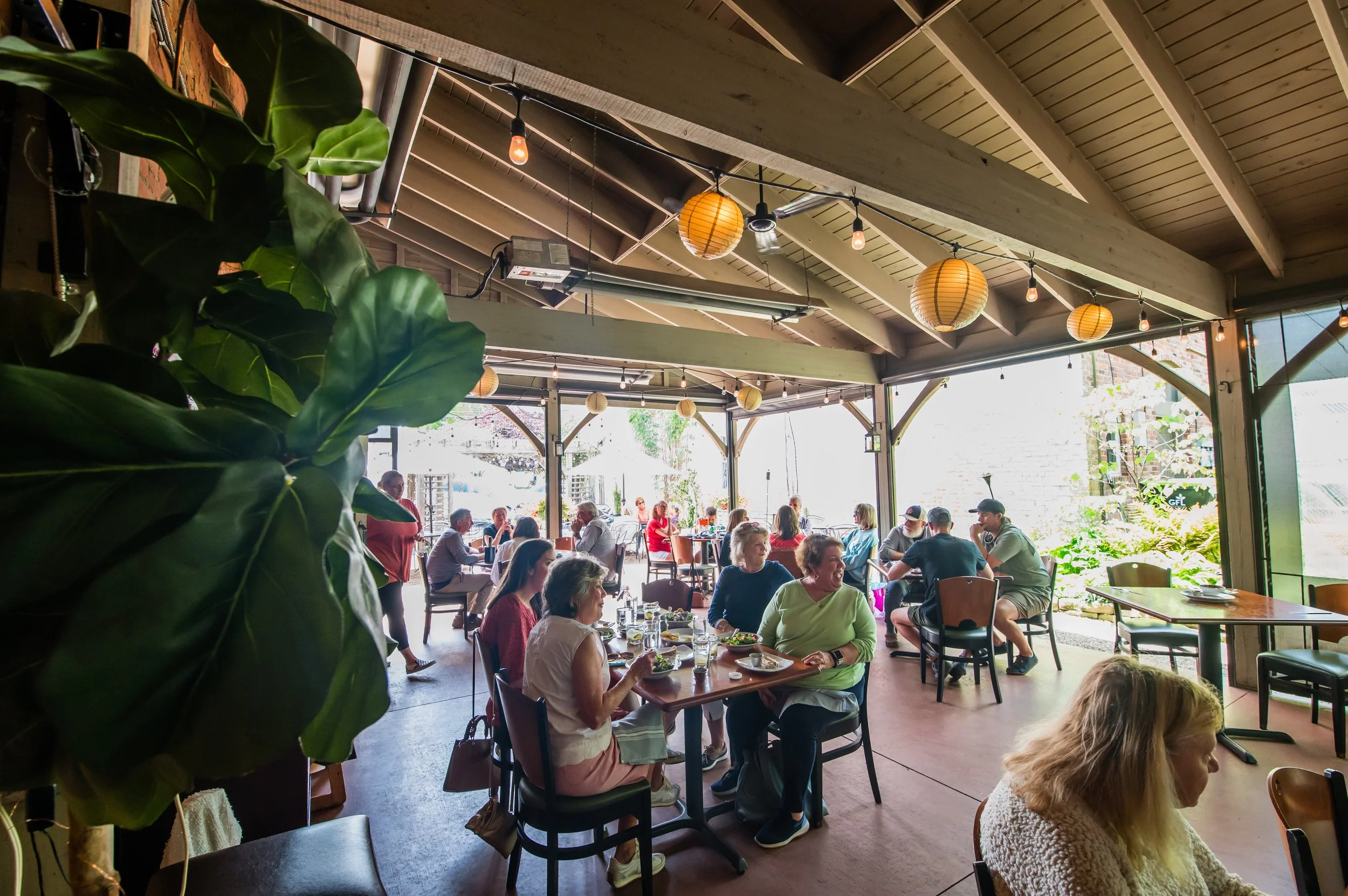 People dining and socializing inside a bright restaurant with large open windows, wooden ceiling beams, and paper lantern and string lights decor.