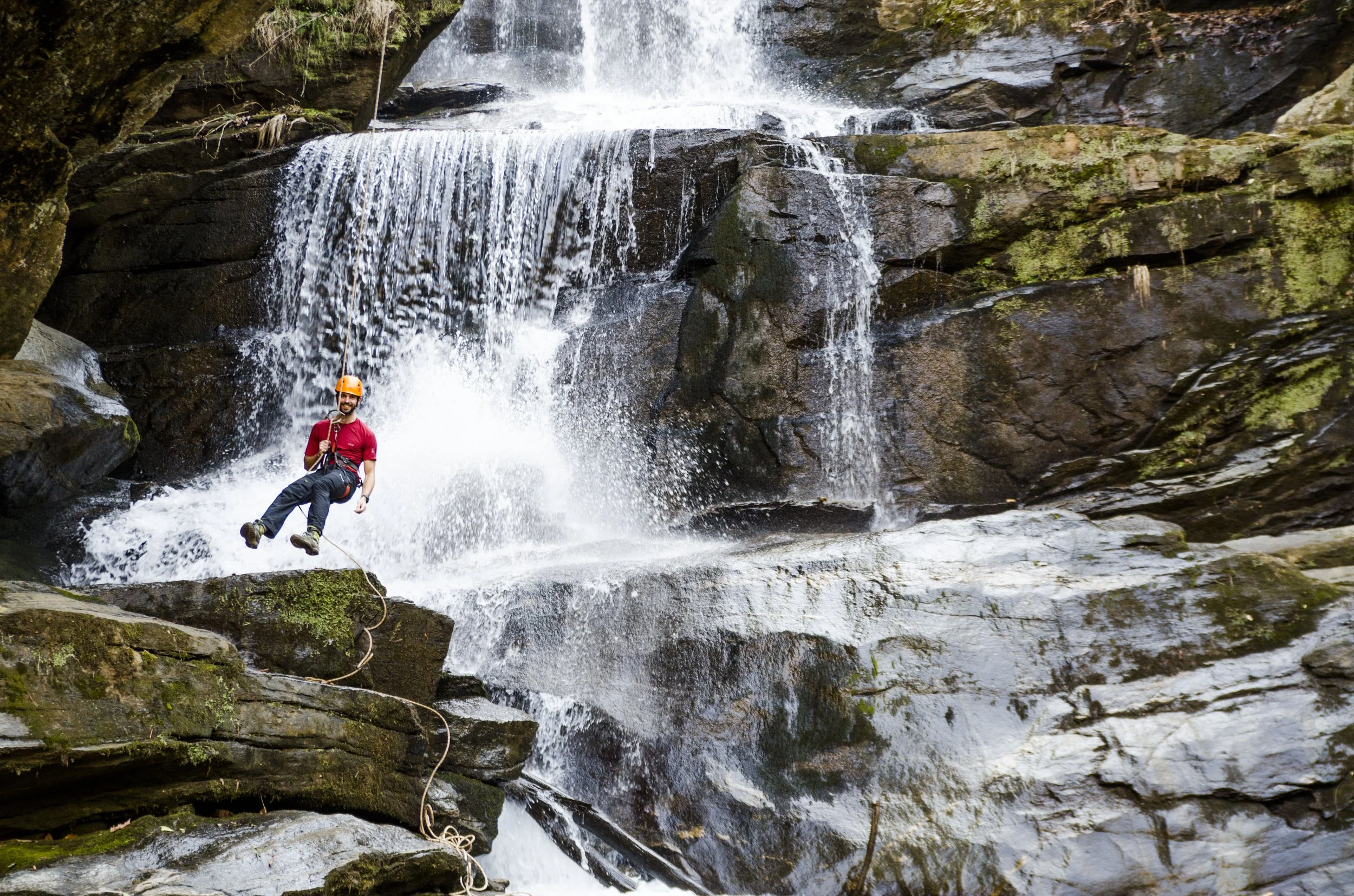 A person wearing a red shirt, black pants, helmet, and harness climbing near a waterfall with water cascading over rocks.