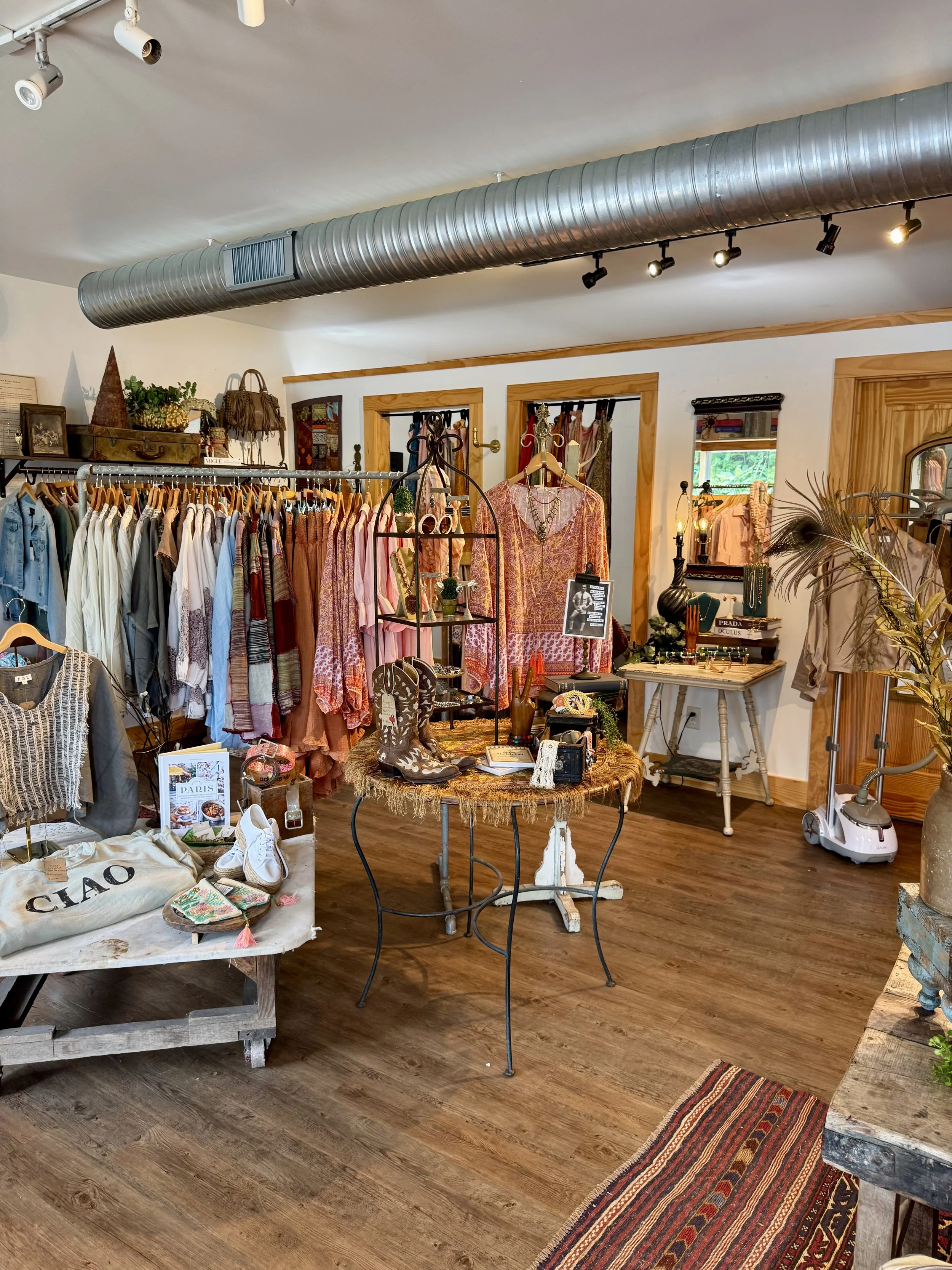 Interior of a boutique clothing store featuring racks of various women's clothing, accessories, and shoes, with a wooden table displaying miscellaneous items, a mirror, and decorative elements, hardwood flooring, and a large industrial air duct on th