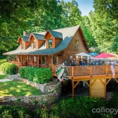 A two-story wooden house with a large deck, umbrellas, and outdoor seating, surrounded by green trees.