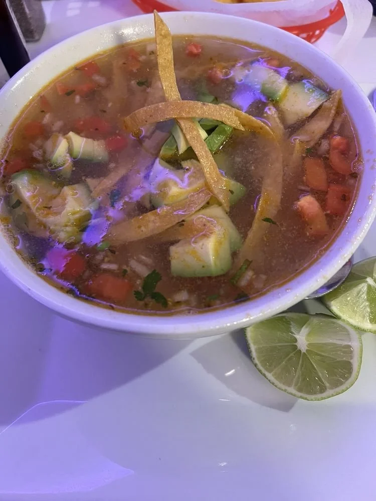 A bowl of hot vegetable soup with visible zucchini, tomatoes, and herbs, garnished with fried tortilla strips; served with lime wedges.