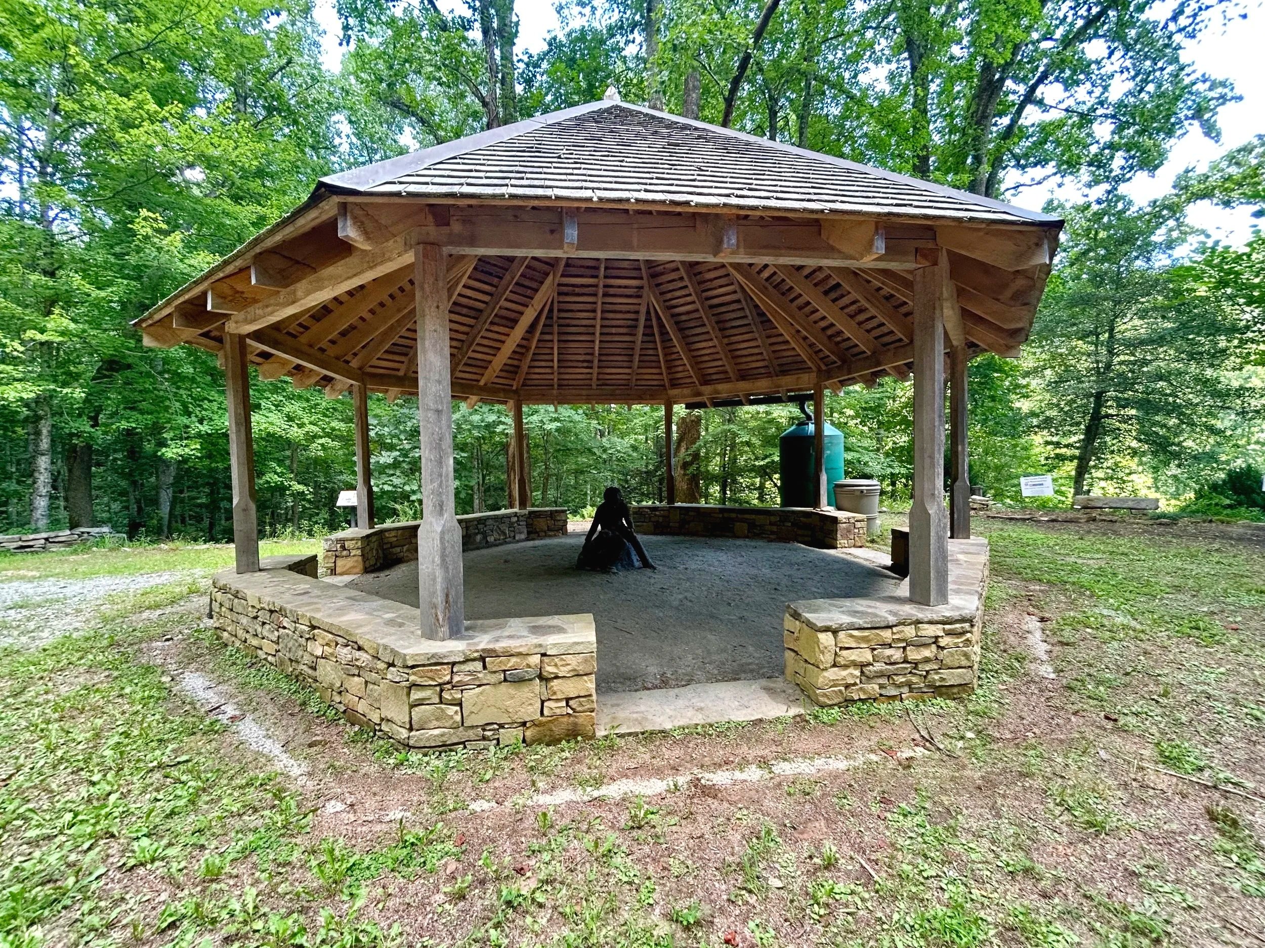 Wooden pavilion with a stone base, surrounded by green trees in a forest setting