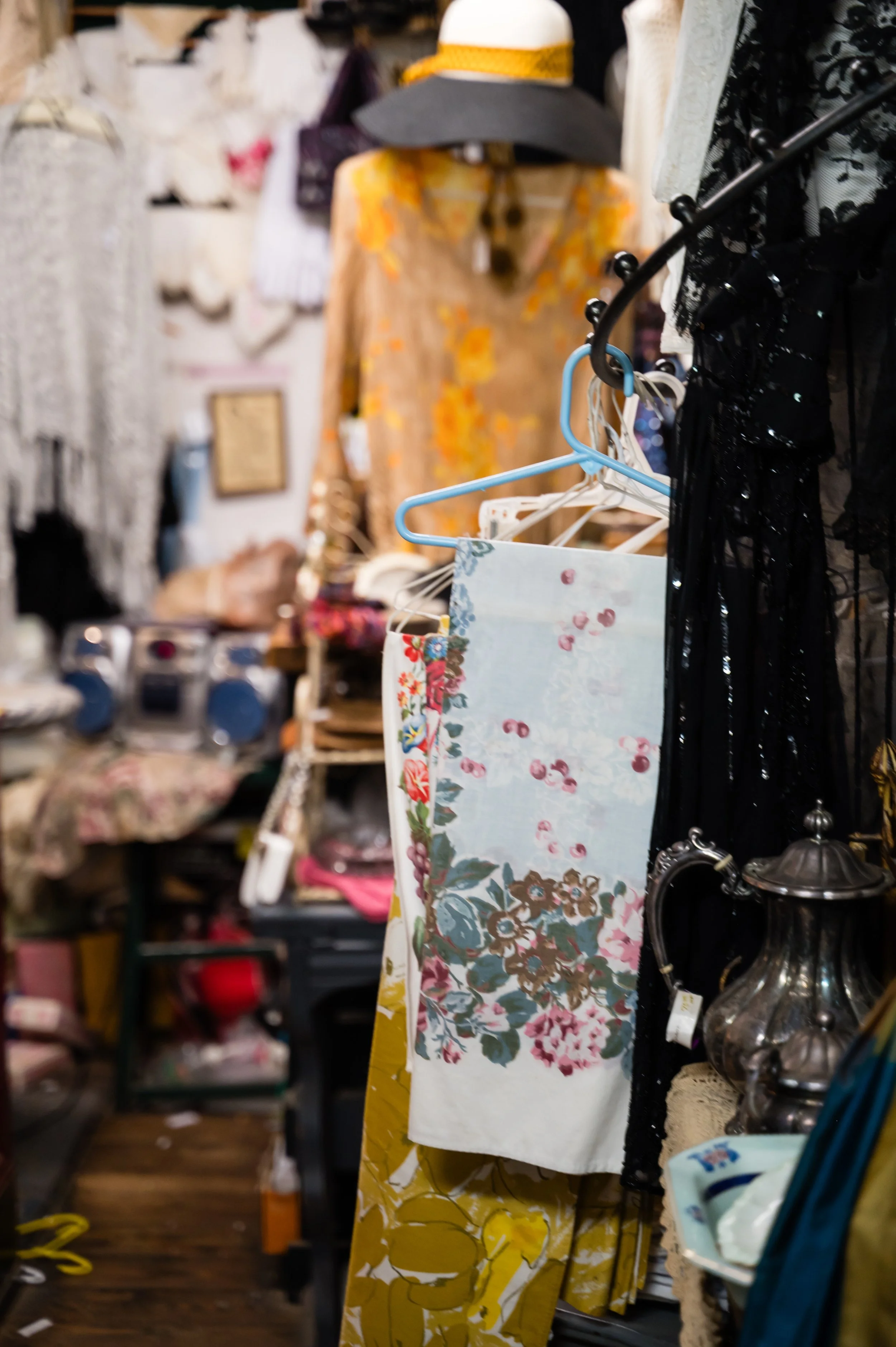 Clothing store display with various garments and accessories, including a floral and yellow patterned dress, shirts, hats, and other items on racks and shelves.
