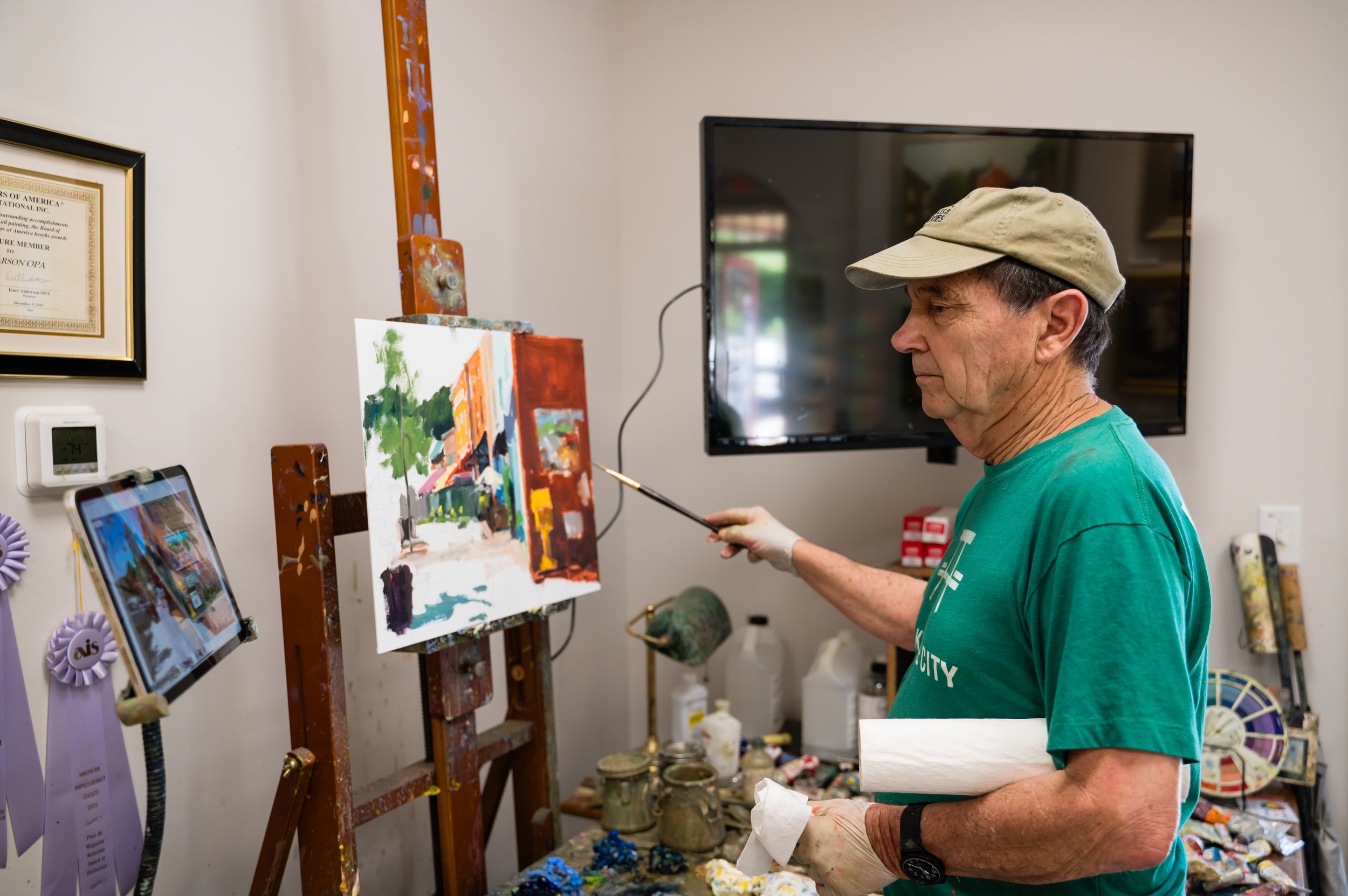 An older man with a beige cap and green T-shirt painting a cityscape on a canvas set up on an easel. He is in a room with art supplies, a television, a framed certificate on the wall, and a tablet.