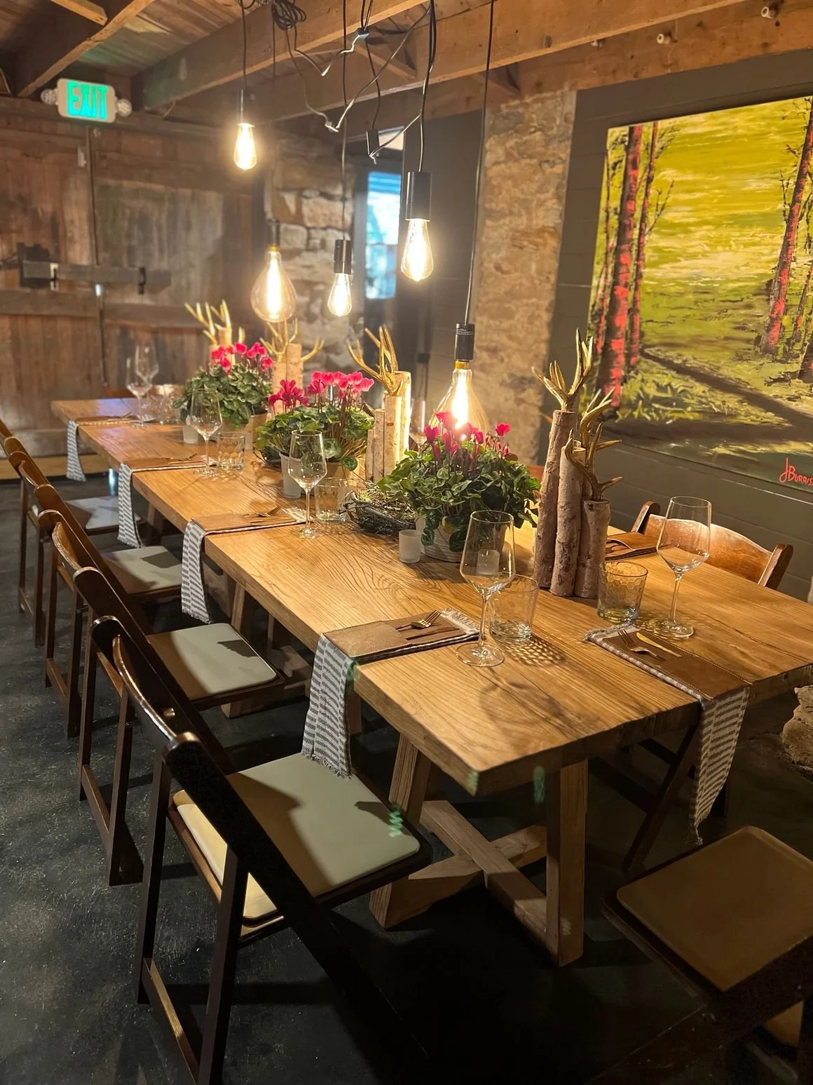 Interior of a rustic dining room with a long wooden table decorated with flowers, small logs, and candles, set for a meal with glasses, forks, and napkins, hanging Edison-style light bulbs, stone and wood walls, and a landscape painting on the wall.