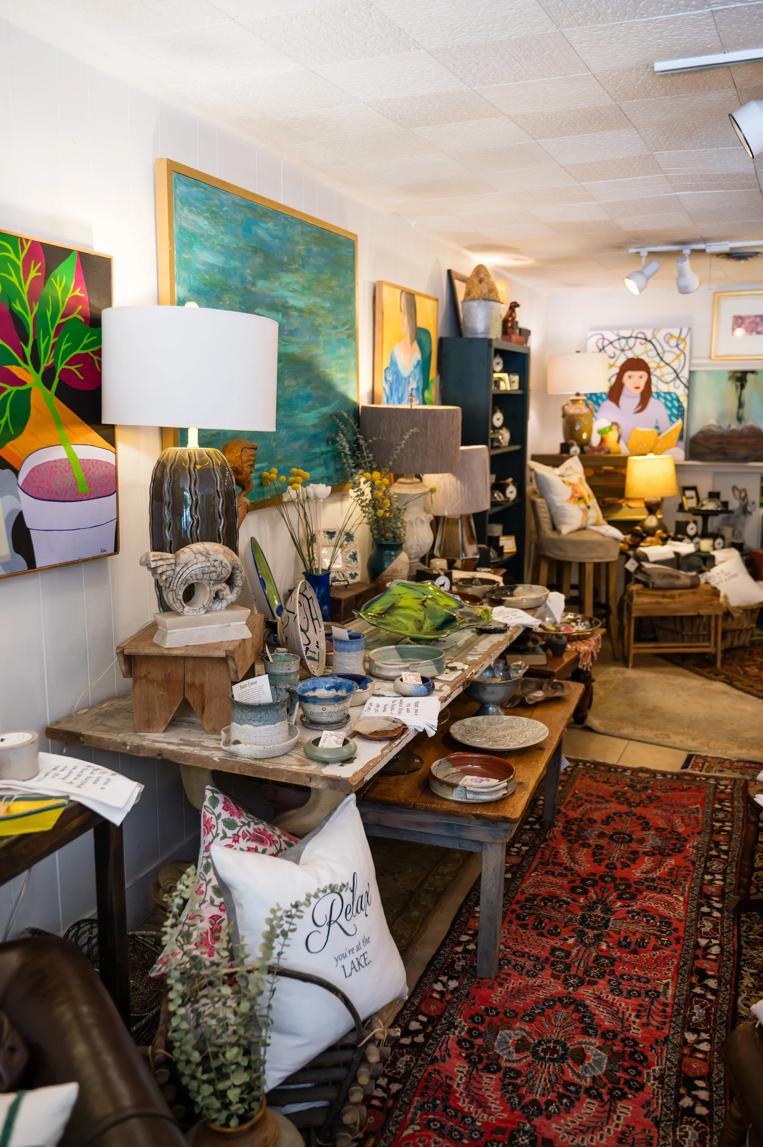 An interior view of an art and home decor store with paintings on the wall, lamps, vases, pottery, and other decorative items displayed on tables and shelves, with a Persian-style rug on the floor.