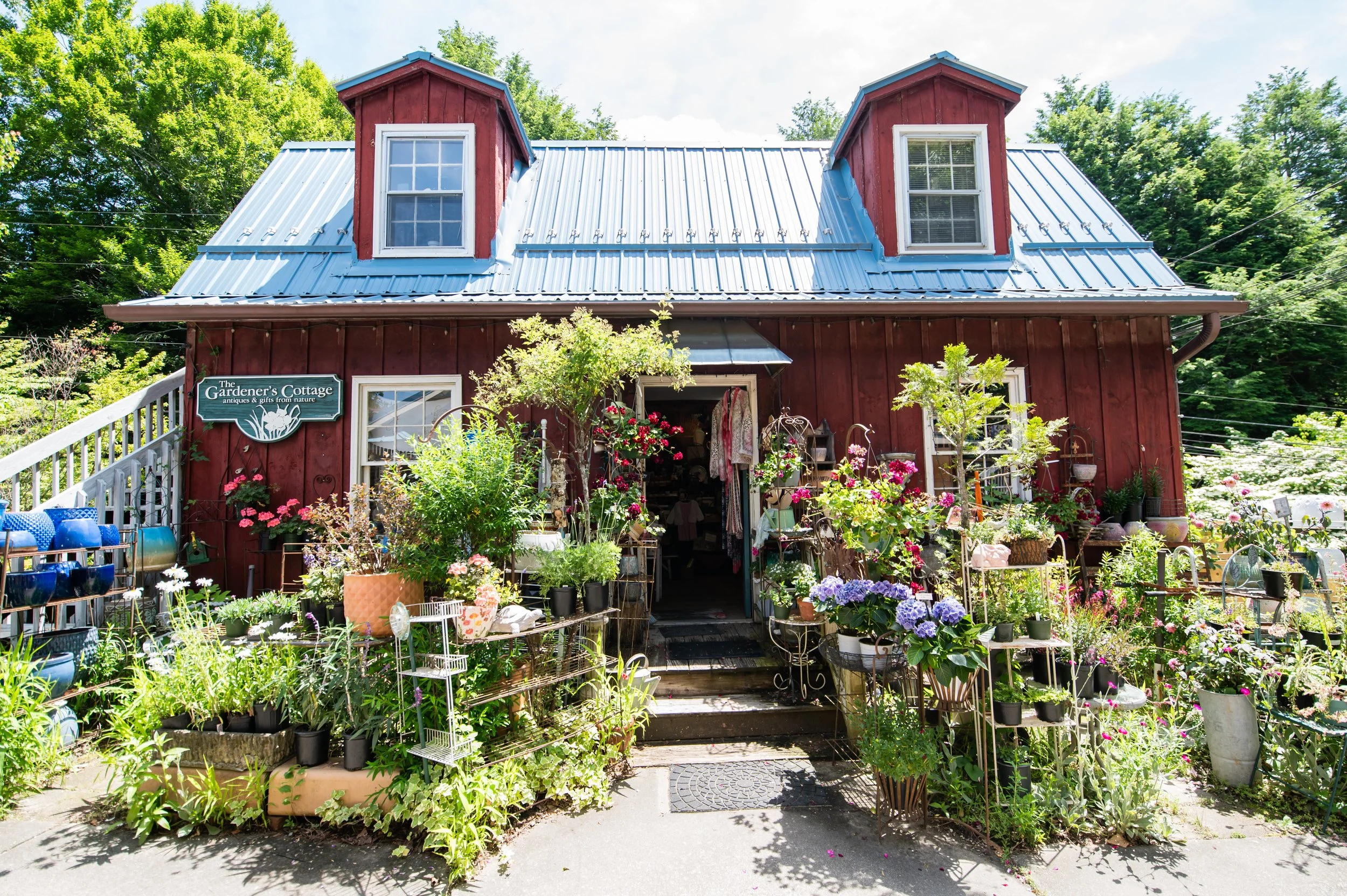 A red barn-style building with a metal roof and two dormer windows, serving as an antiques and gift shop called "The Gardener’s Cottage." The shop is surrounded by colorful potted plants, flowers, and garden decorations outside.