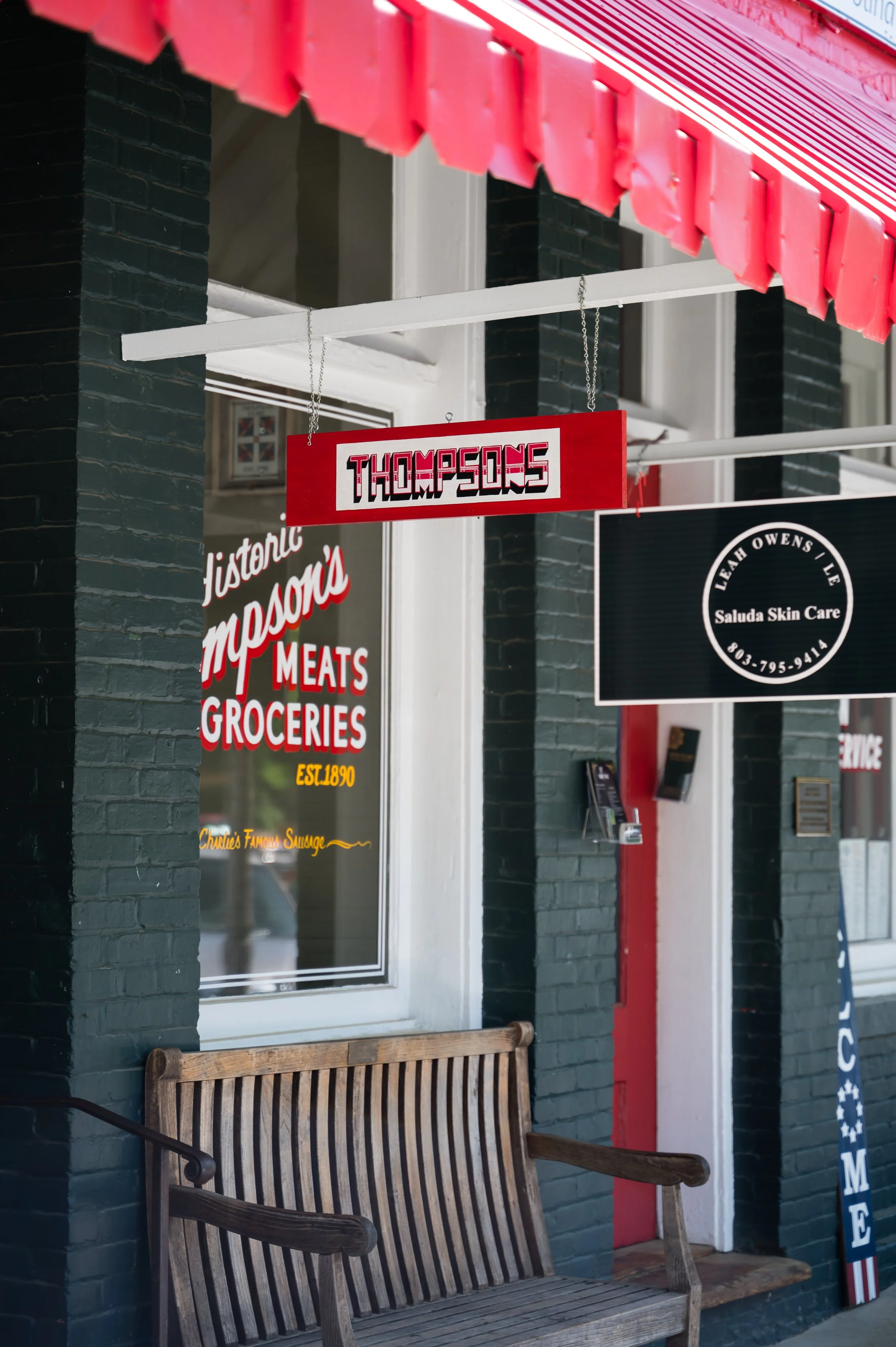Storefront with black brick exterior, red awning, hanging sign with 'Thompsons', and a wooden bench outside.
