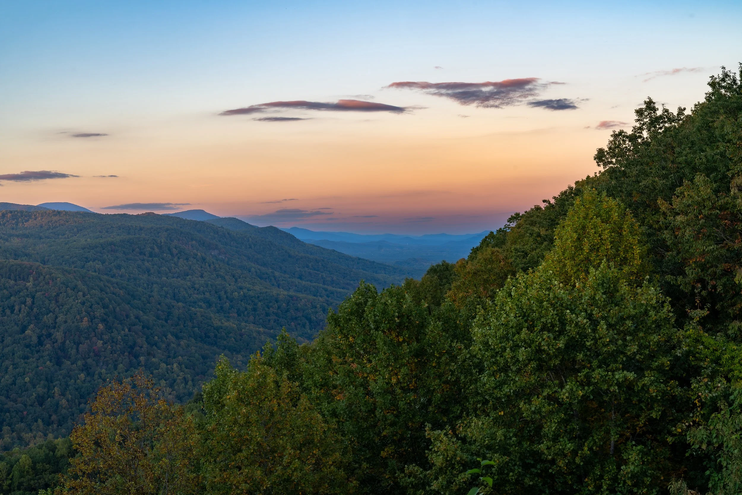 A scenic view of a mountain range during sunset with a sky transitioning from light blue to soft pink and peach hues, and dense green trees in the foreground.