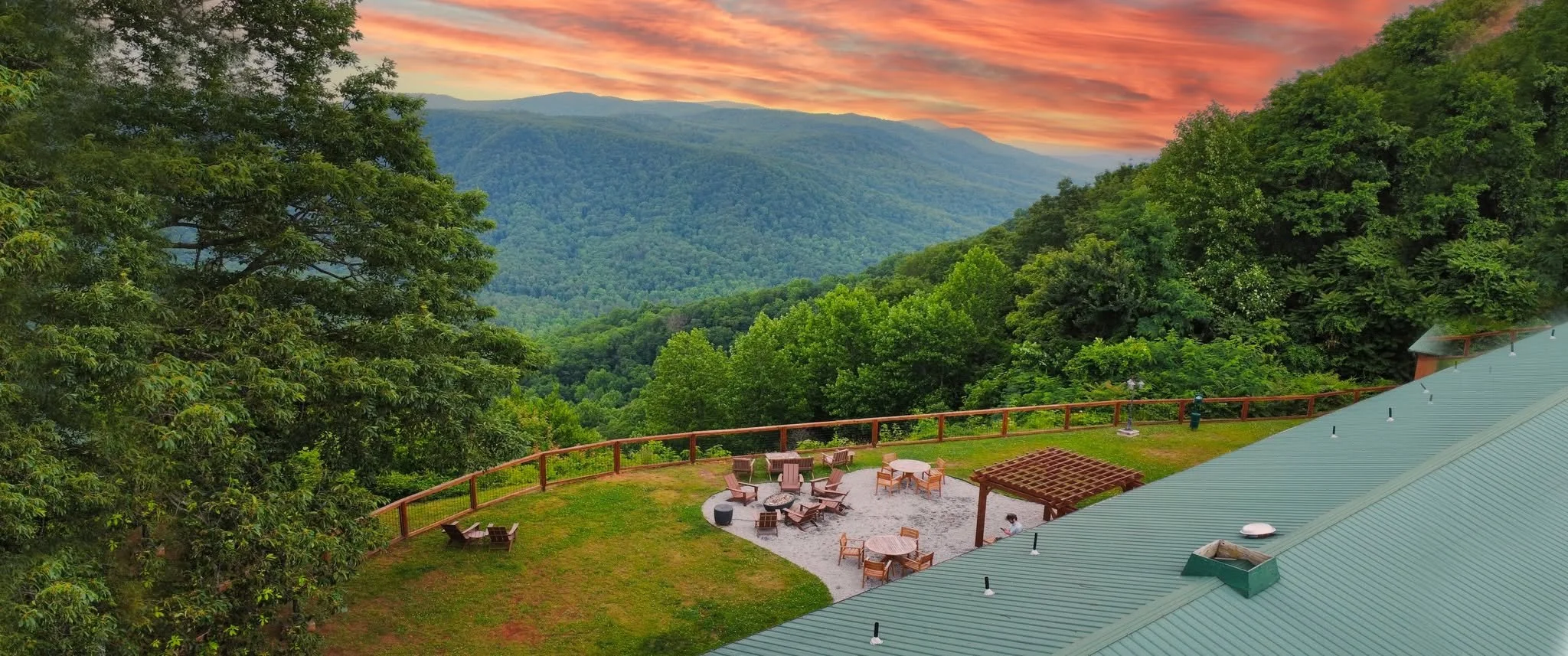 A scenic view of a mountain landscape during sunset, with dense green forest and a firepit area with chairs in the foreground, adjacent to a building with a green metal roof.
