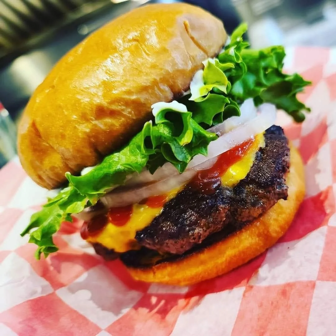 Close-up of a cheeseburger with a beef patty, lettuce, onion, ketchup, mustard, and a toasted bun on red checkered paper.
