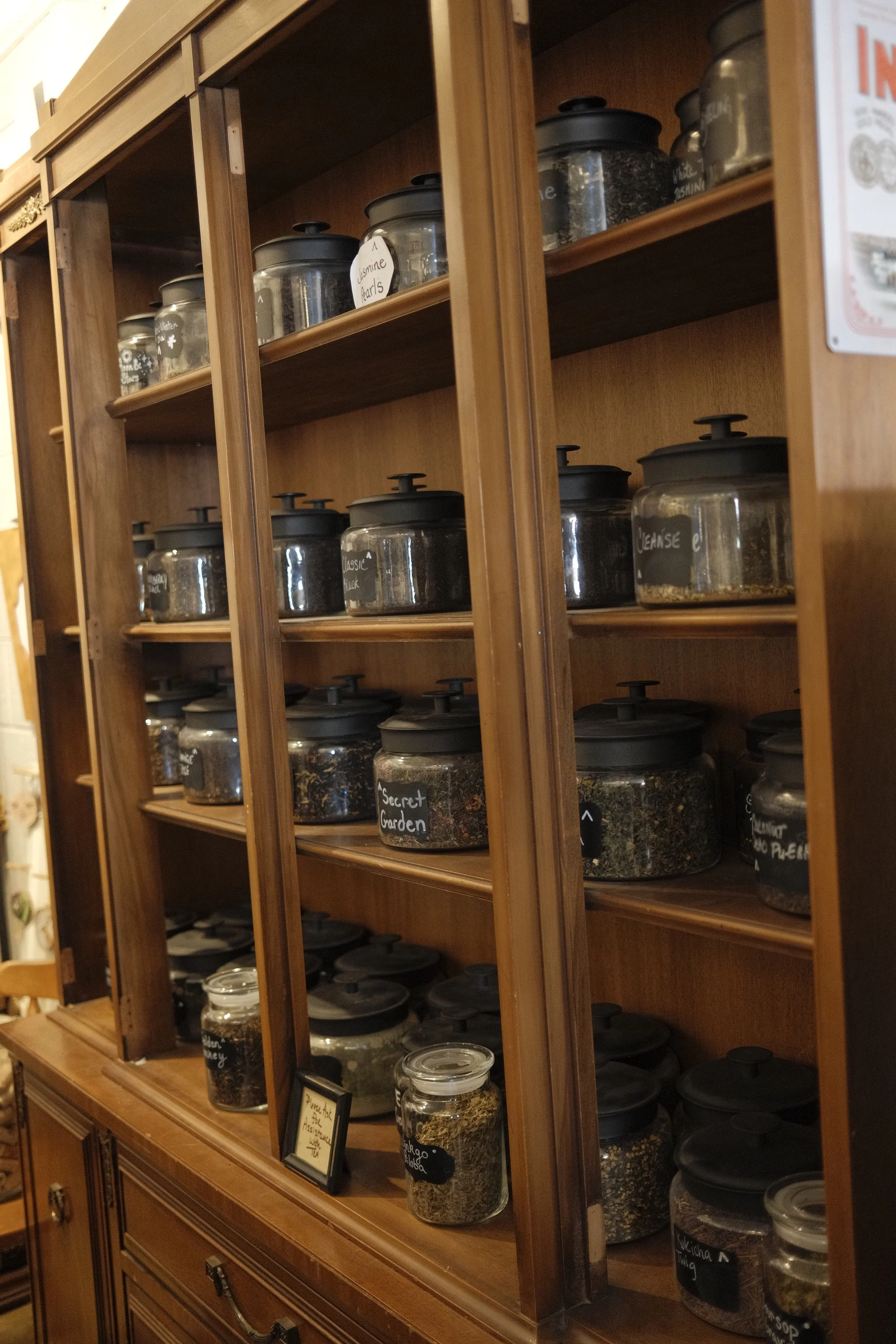 Wooden shelf with glass jars containing various herbs, spices, and dried ingredients, each labeled with small handwritten tags.