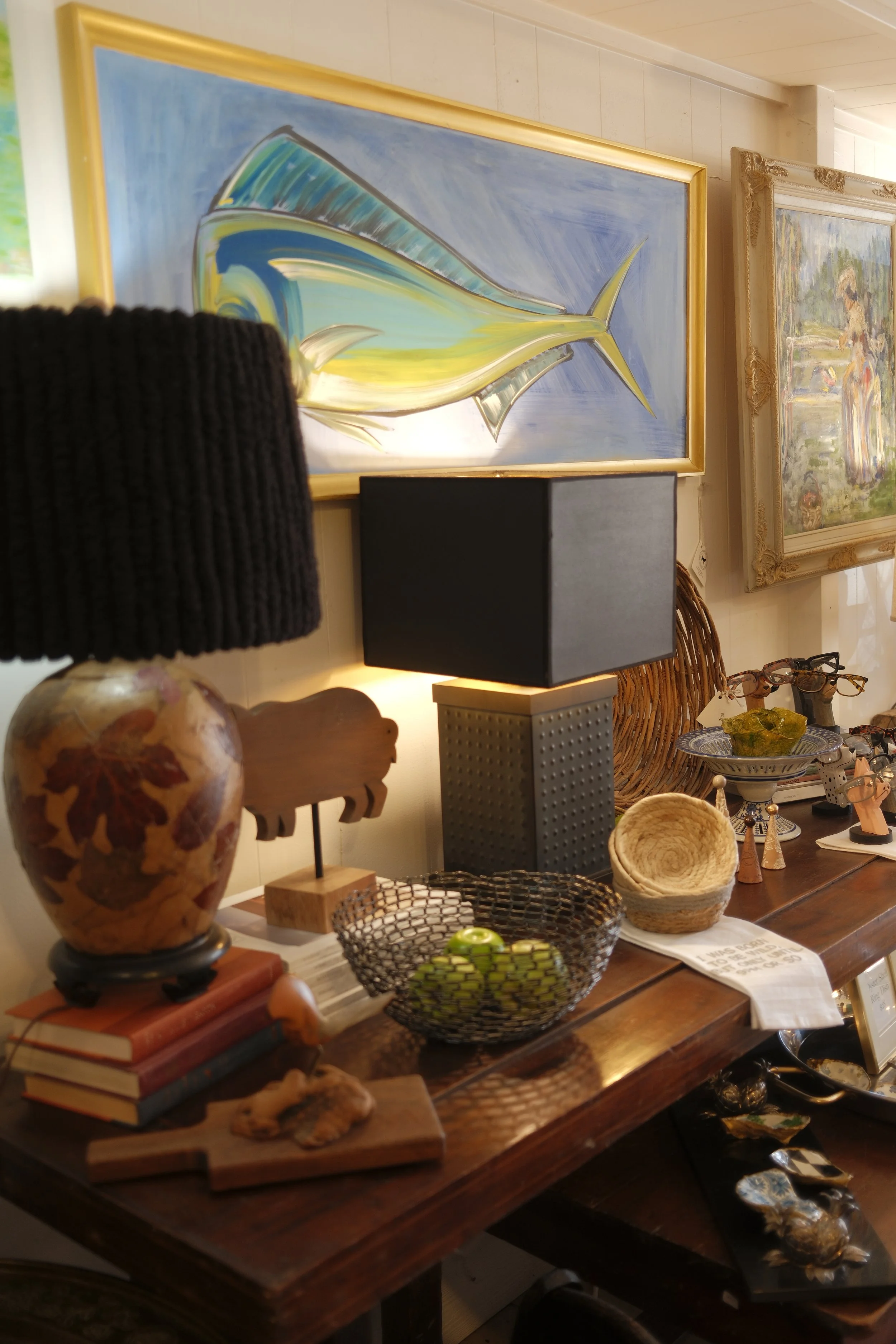 An interior display of various decorative objects including a colorful abstract painting of a fish, a black lampshade on a table lamp, stacked books, a ceramic vase with floral patterns, a woven basket, a small wooden sign, and other small decorative