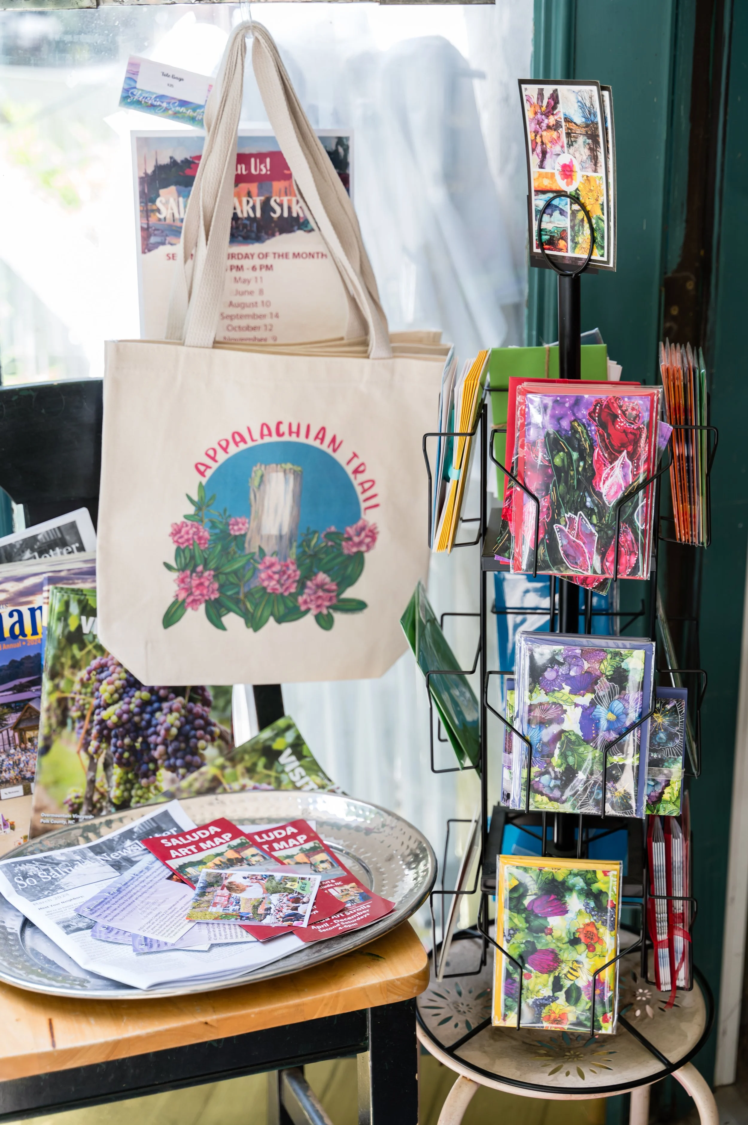 A display stand with colorful artwork prints, a tote bag with an Appalachian Trail logo, and pamphlets for a local art event. Newspapers and flyers are on a round table nearby.