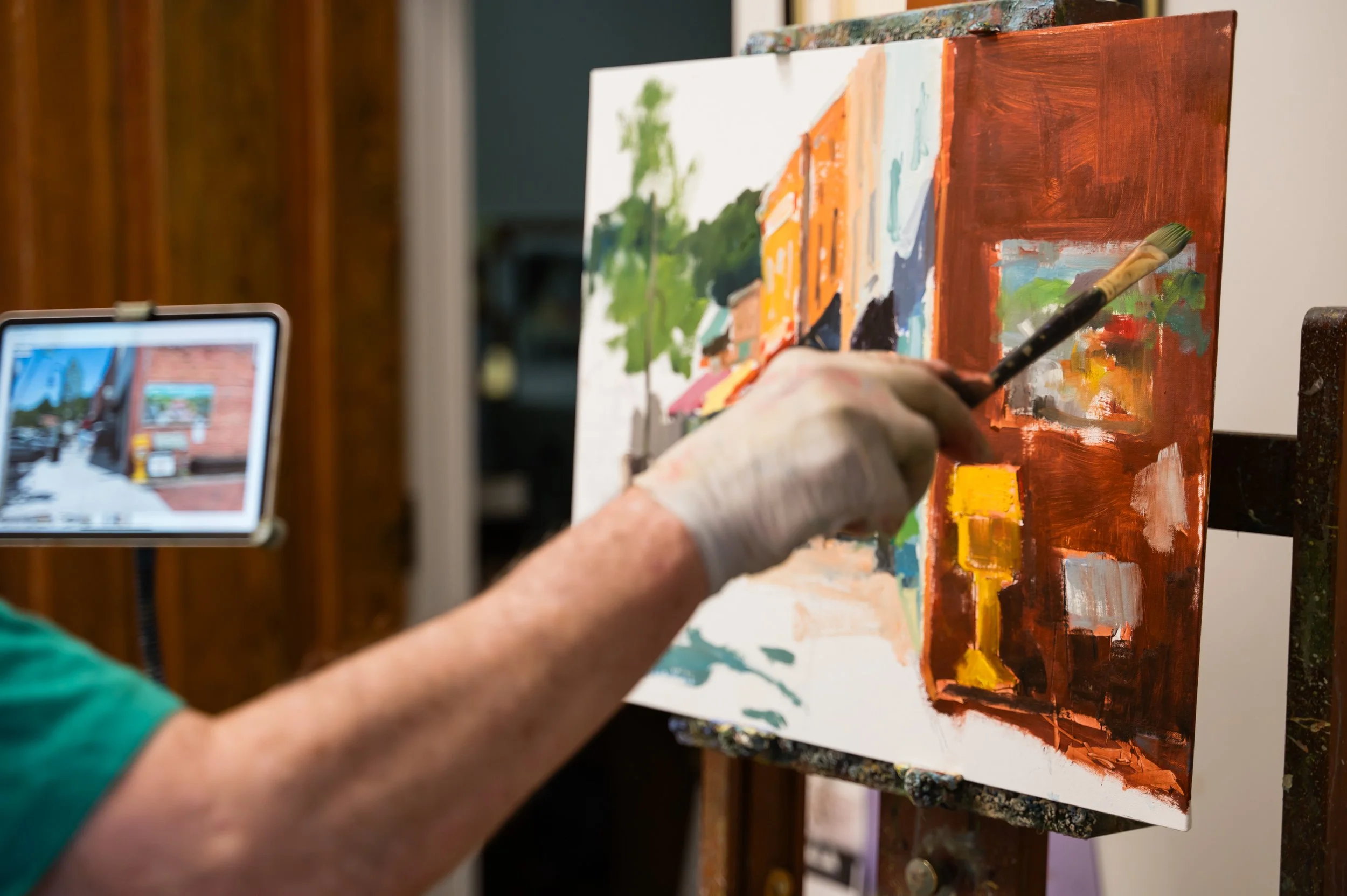 Close-up of an artist's hand holding a paintbrush and painting on a canvas with an abstract landscape, with a tablet on a stand in the background displaying a reference image.
