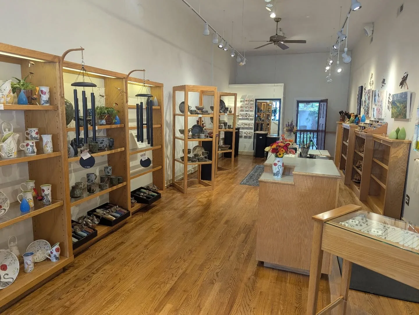 Interior of an art and craft store with wooden shelves displaying pottery, jewelry, and decorative items, a central display table with a flower vase, artwork on the walls, wood flooring, and track lighting.