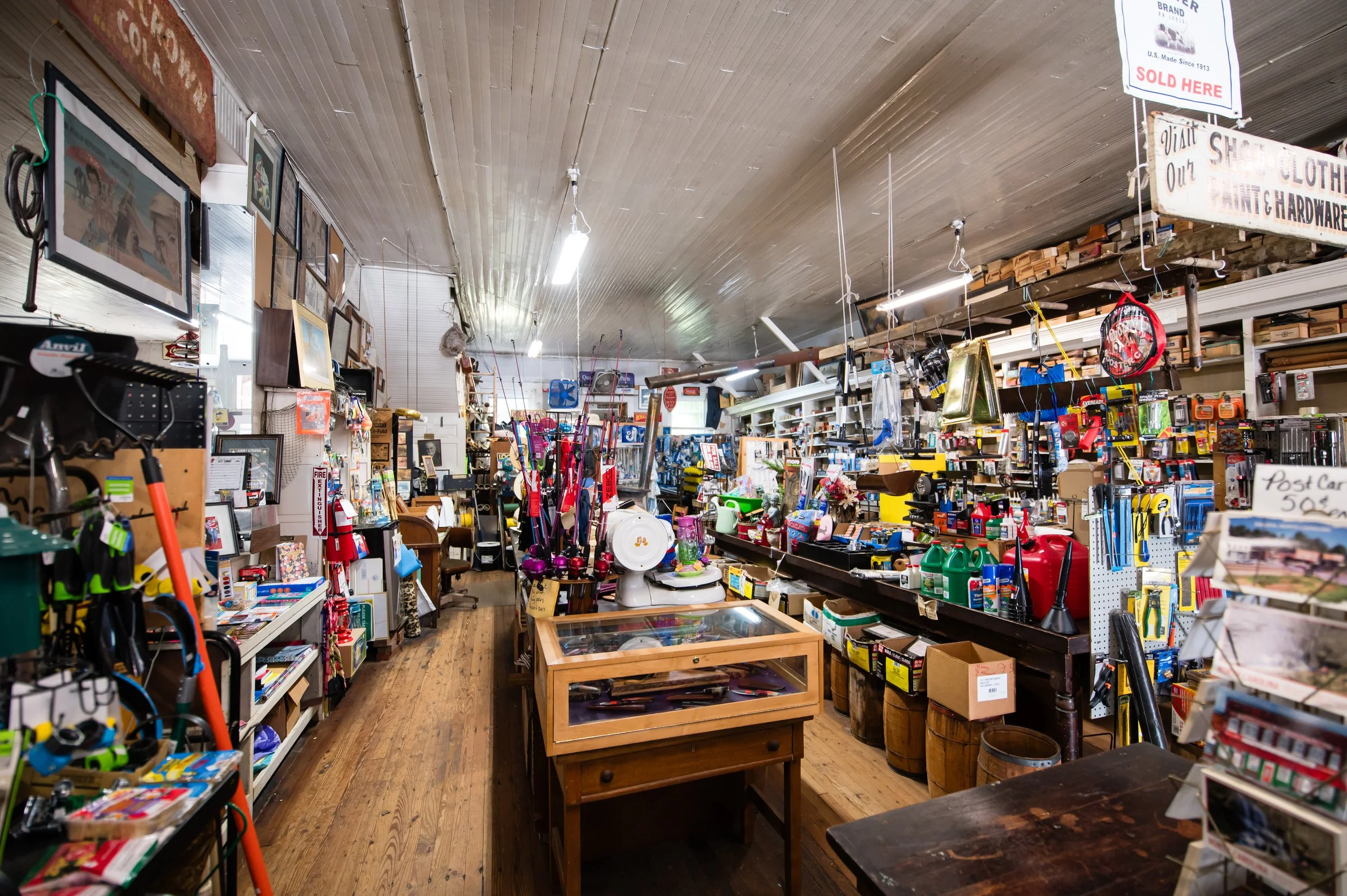 Inside a thrift or antique store with shelves and tables filled with various used items such as picture frames, household goods, tools, and decorative objects.