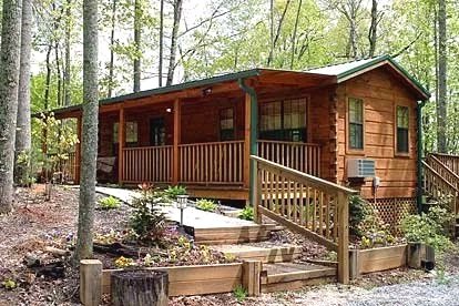 A wooden cabin with a porch, set amidst trees in a forest, with a small garden and stairs leading up to the entrance.