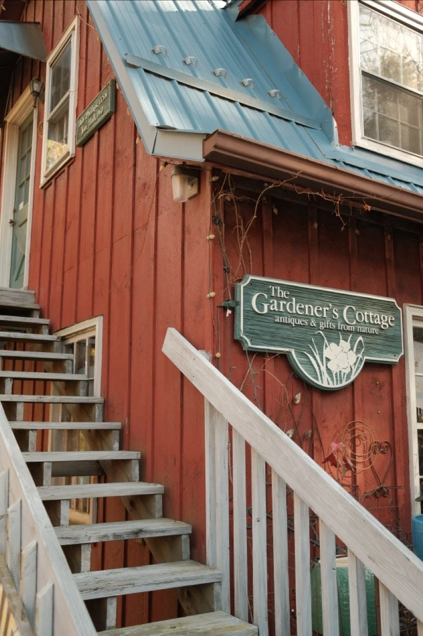 Exterior of a red wooden building with a wooden staircase leading up to a door. There is a green sign that reads 'The Gardener's Cottage antiques & gifts from nature' and features illustrations of flowers and gardening tools.