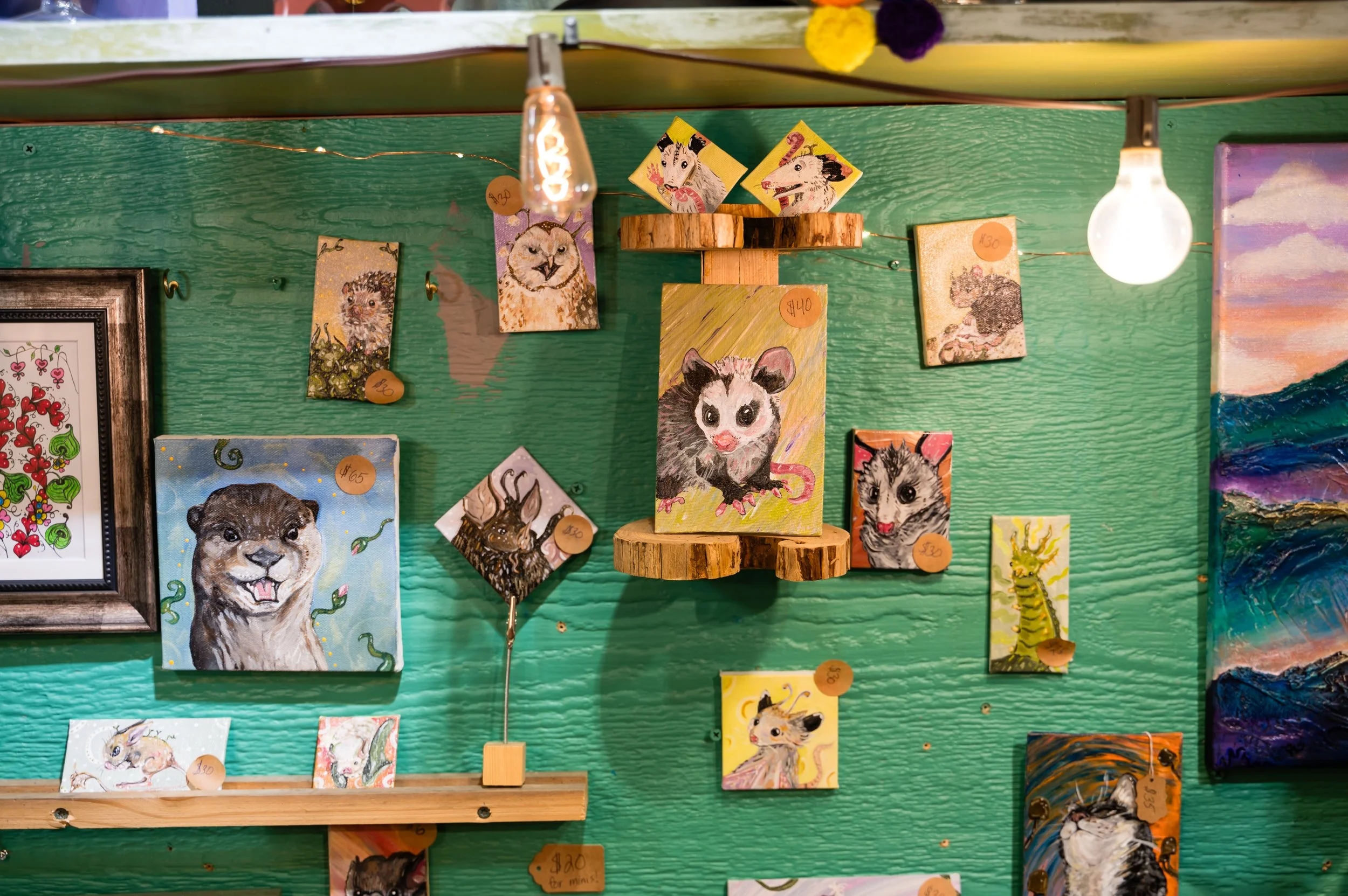 A green wall decorated with various illustrated animal art pieces, including lions, wolves, hamsters, and a hedgehog. Some artworks are framed, others are on small canvases, with price tags attached.