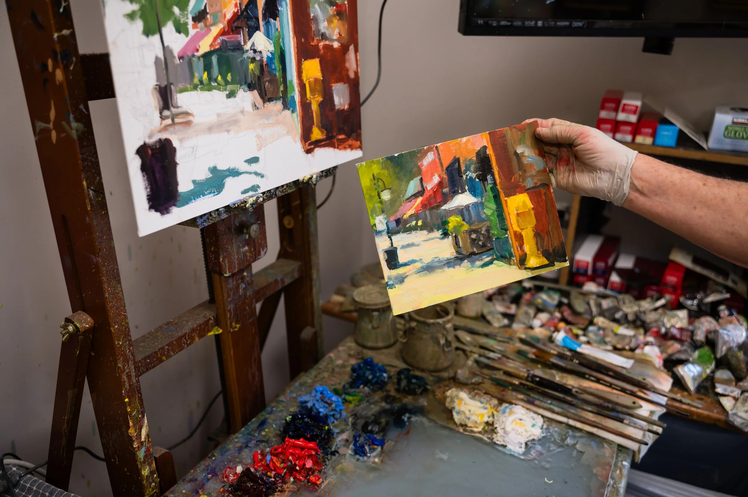 An artist working in a studio, holding a small colorful painting of a city street scene with buildings, trees, and sky. There are paintings in progress, art supplies, and a cluttered workspace in the background.