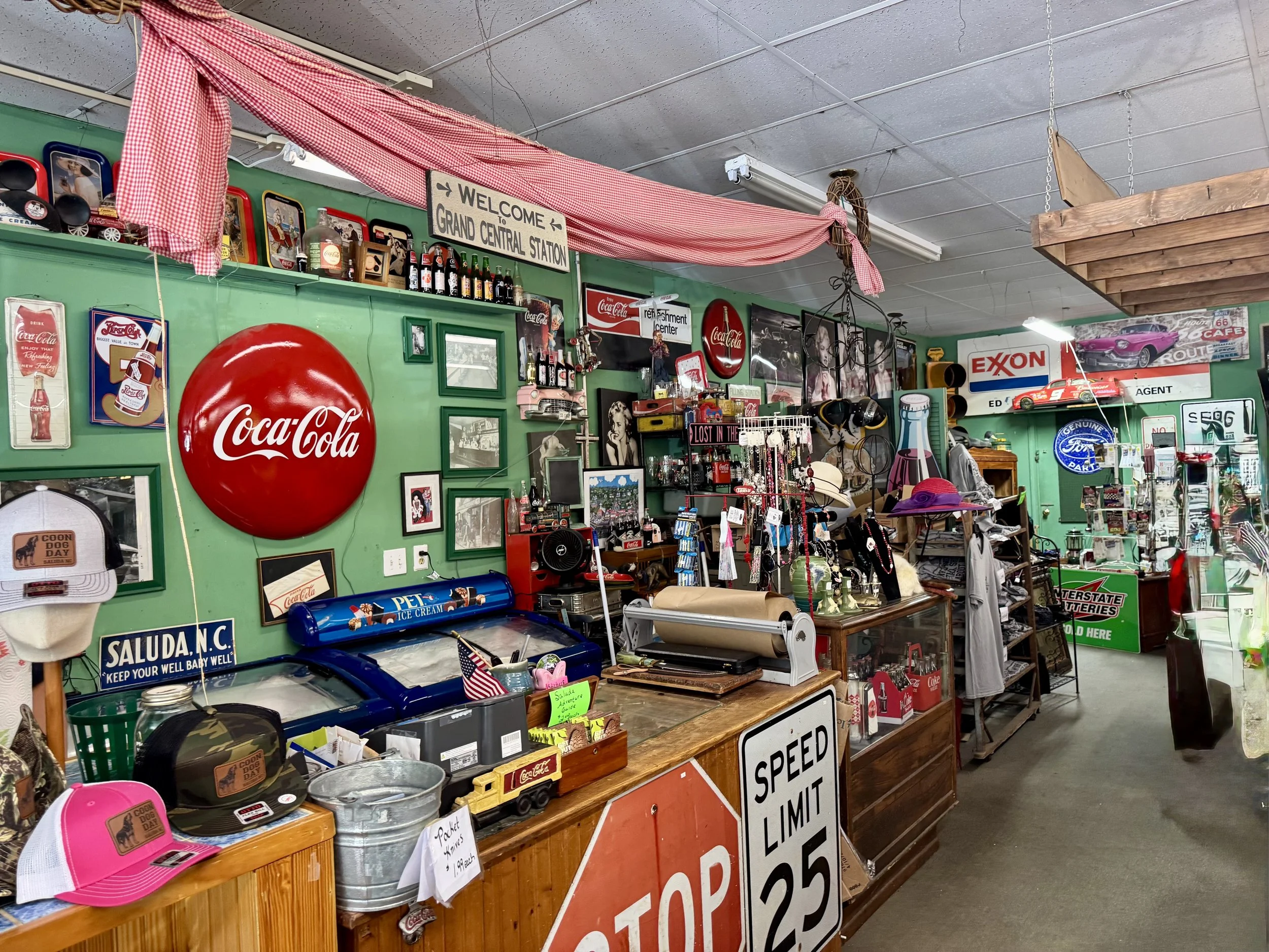 Inside a vintage thrift store with memorabilia and collectibles including Coca-Cola signs, vintage toys, hats, and signs, with a sign welcoming to Grand Central Station and other vintage advertisements.