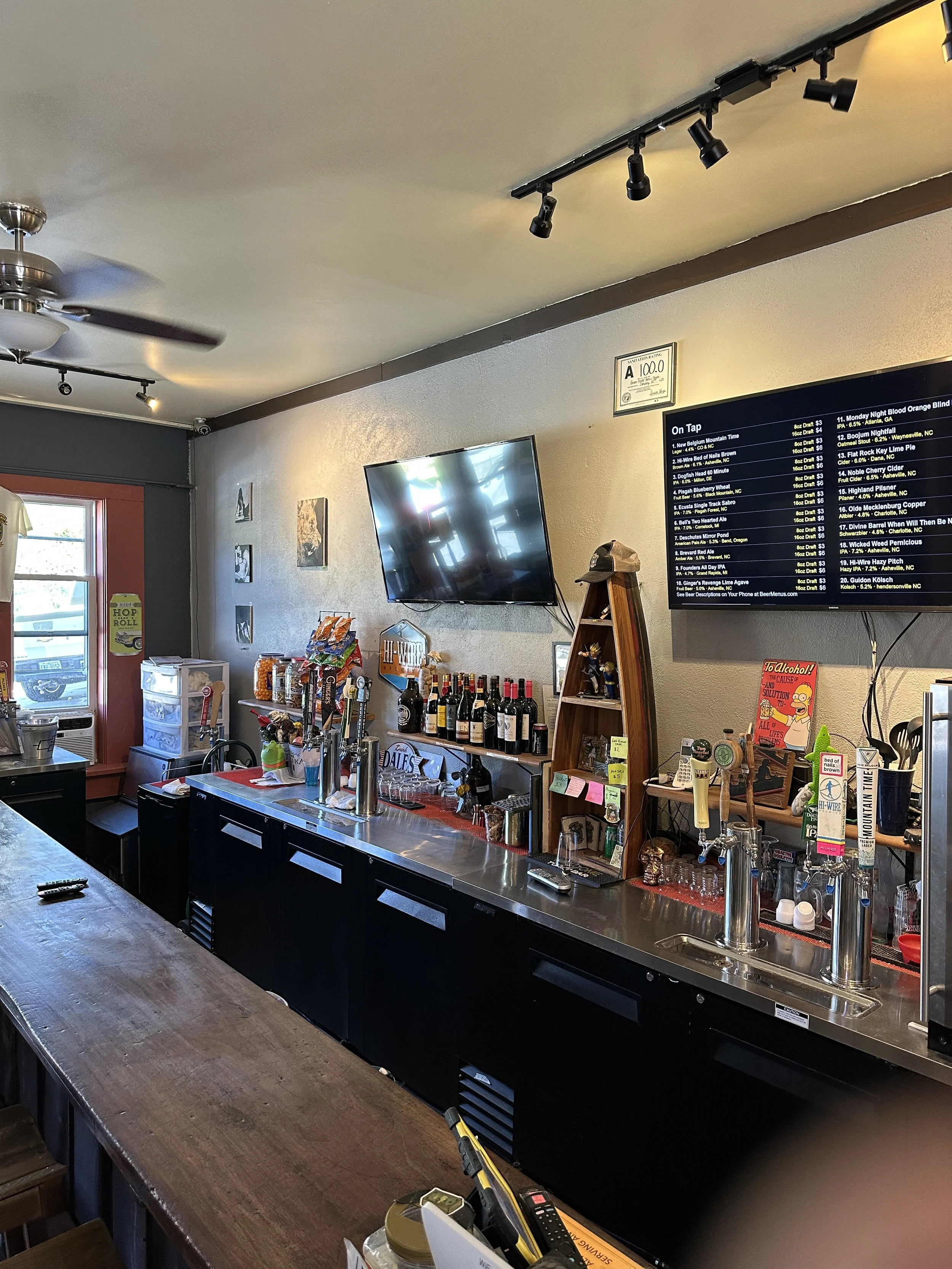 Interior of a bar or restaurant with a wooden counter, shelves with wine bottles, a digital menu board, and a TV on the wall. There are some snacks and bar tools on the counter.