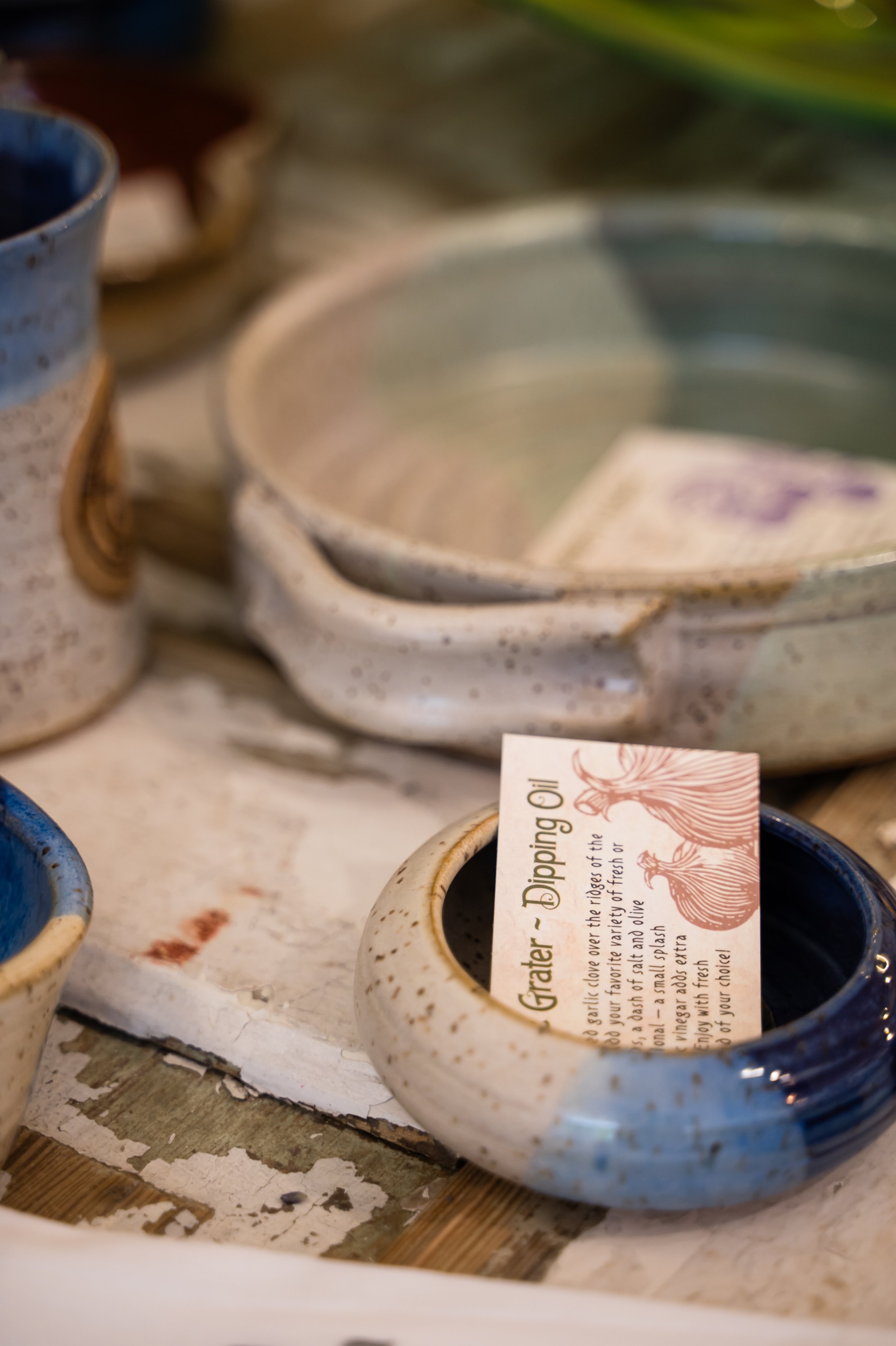 Assorted ceramic bowls and cups on a rustic wooden table, with a label inside one of the bowls mentioning herbs and garlic.