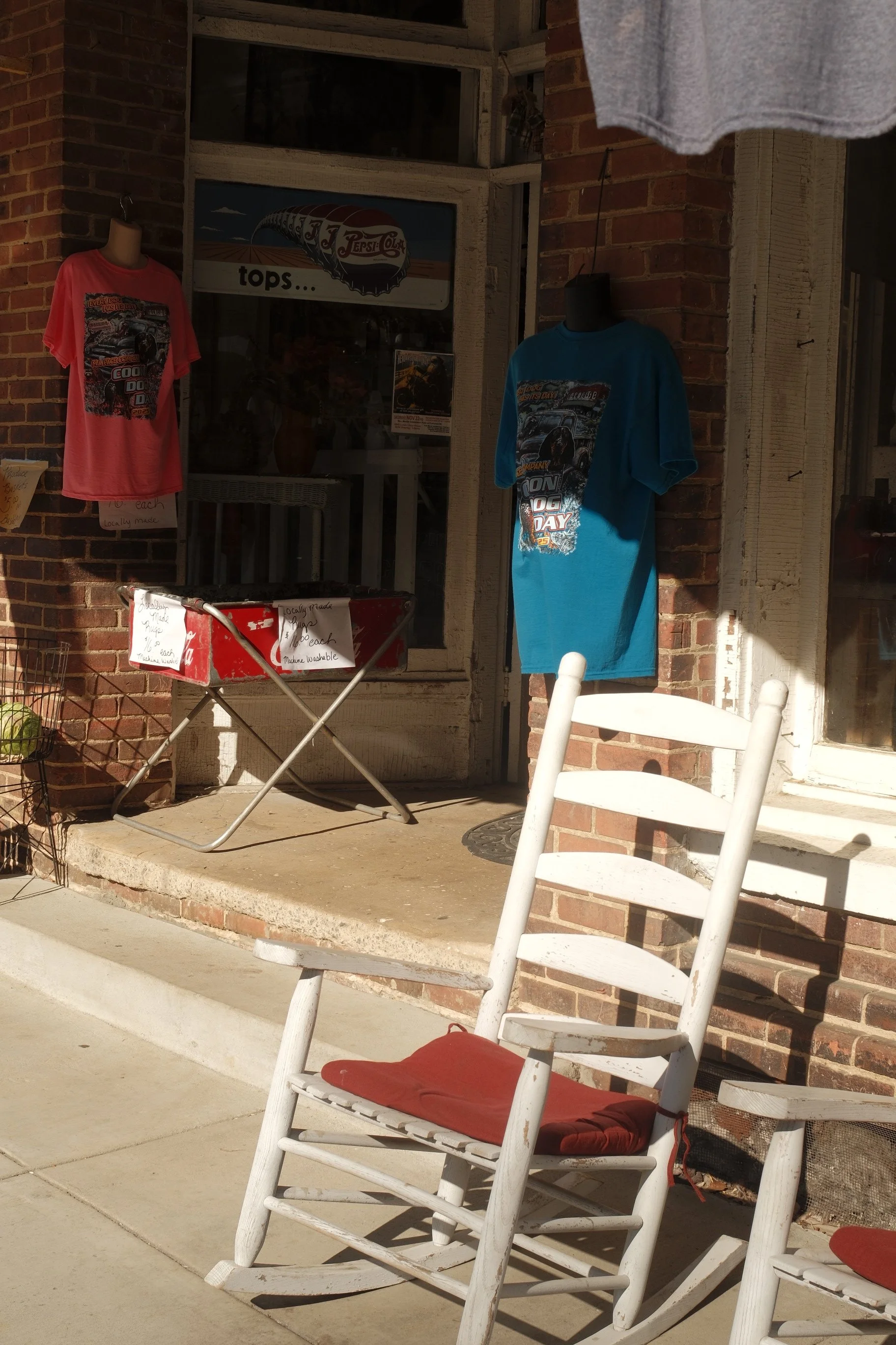 A porch scene with two white rocking chairs with red cushions, a folding table with signs and merchandise, and two mannequins wearing colorful t-shirts in front of a brick building with a window and an American flag-themed sign.