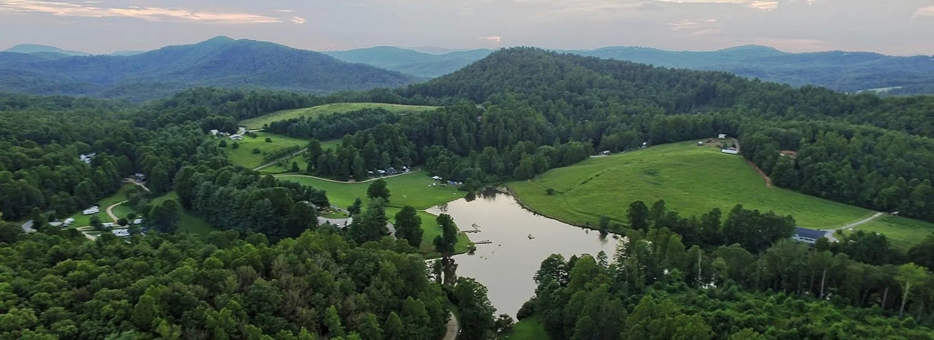 A scenic aerial view of a lush green landscape with rolling hills, a small lake, a forested area, and a few scattered buildings, set against a backdrop of distant mountains and a cloudy sky.
