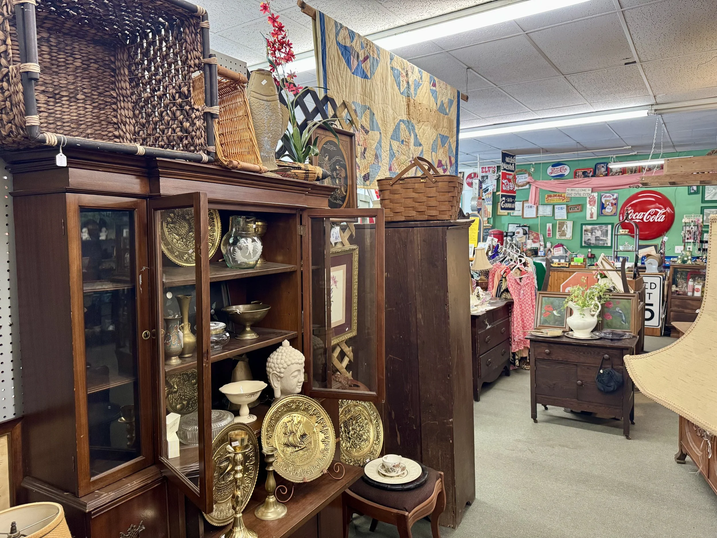 An indoor thrift store or antique shop with various vintage items, including wooden furniture, decorative plates, and metal trays. There are hanging quilts and signs in the background, suggesting a eclectic collection of secondhand artifacts.
