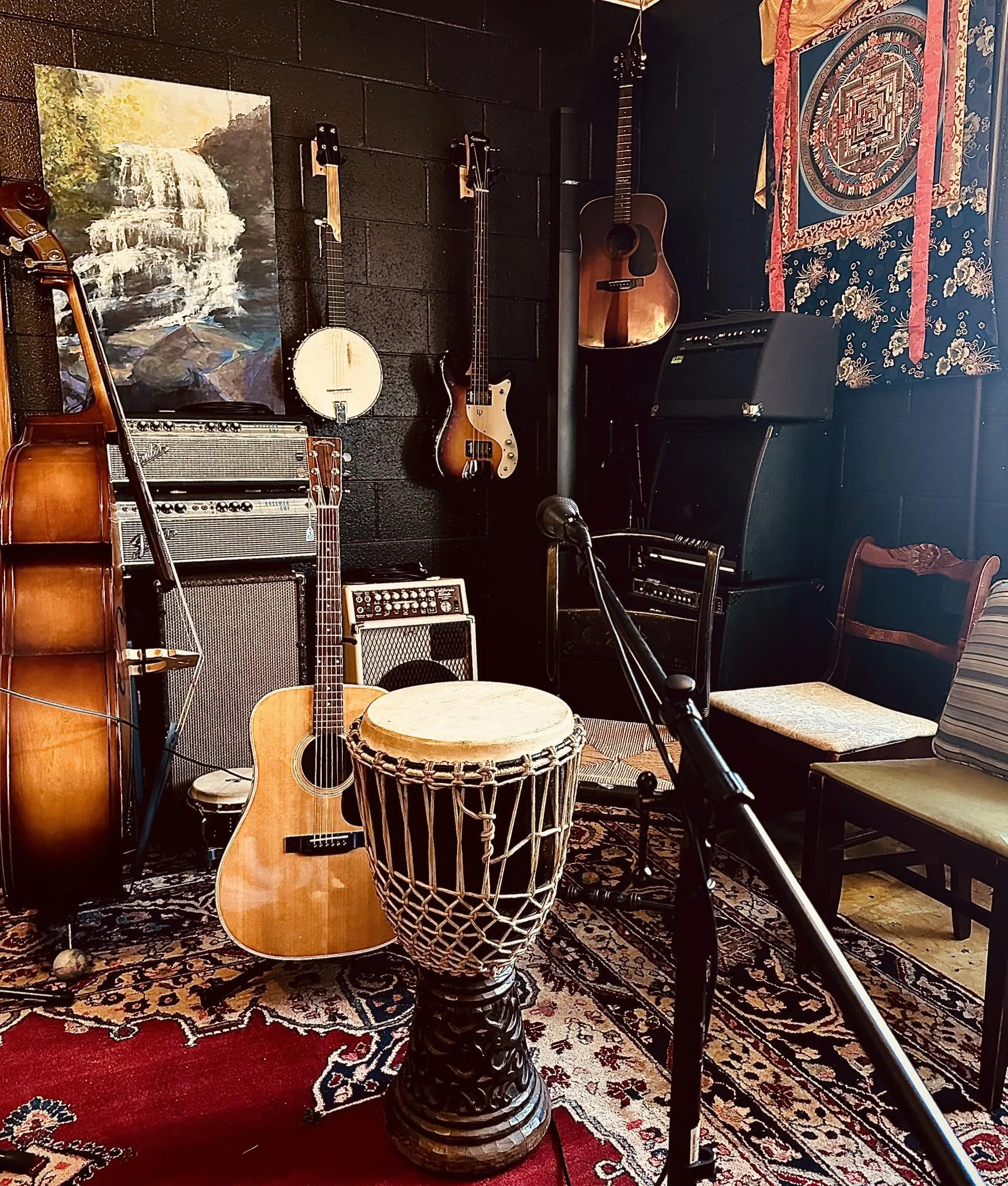 Musical instruments including an acoustic guitar, a bass guitar, and a banjo are arranged against a black wall, with a drum in the foreground. A painting of a waterfall hangs on the left, and a colorful tapestry with geometric and floral patterns is 