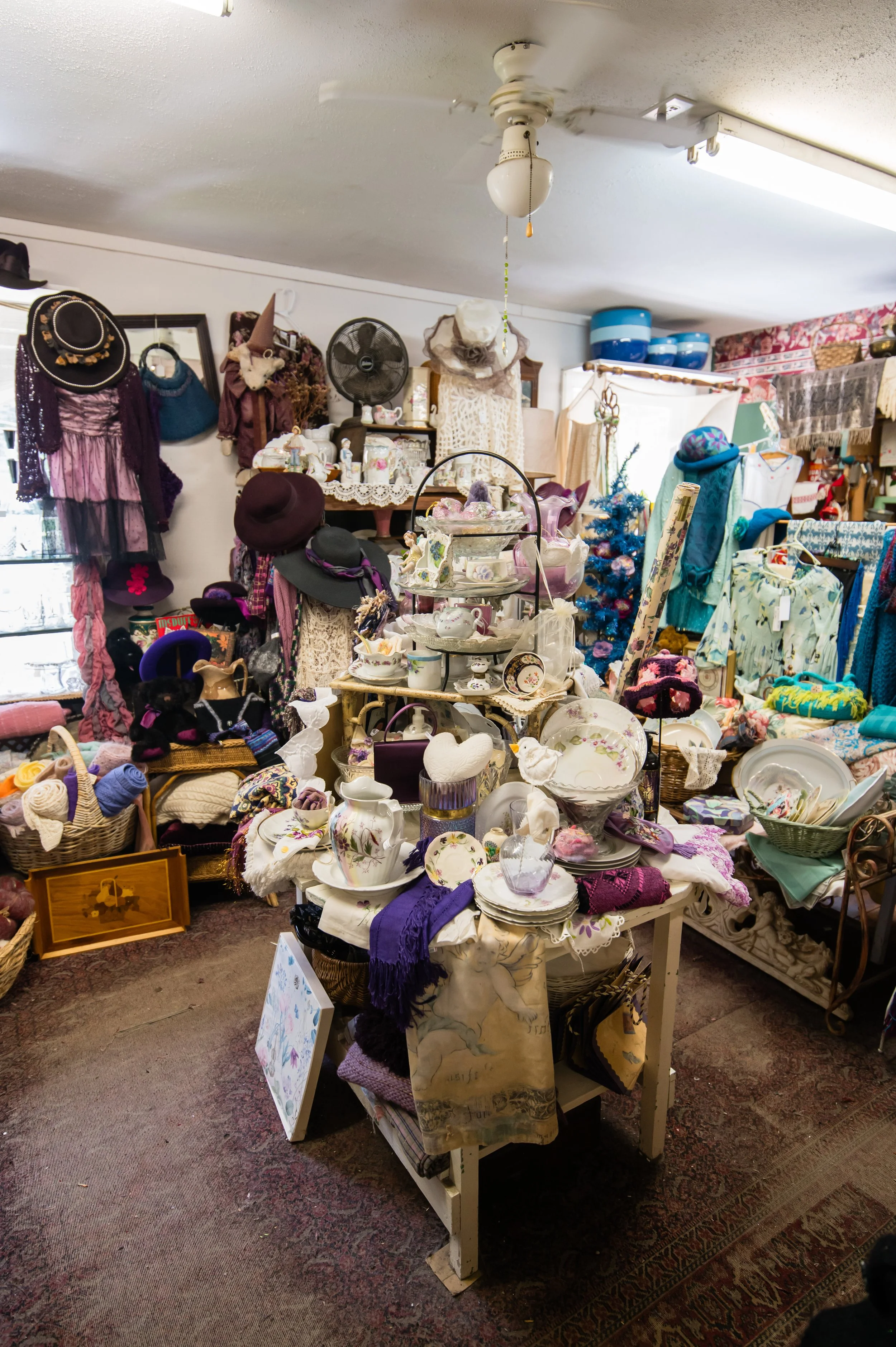 An indoor thrift store display with various hats, dishes, and decorative items on tables and shelves, and a ceiling fan above.