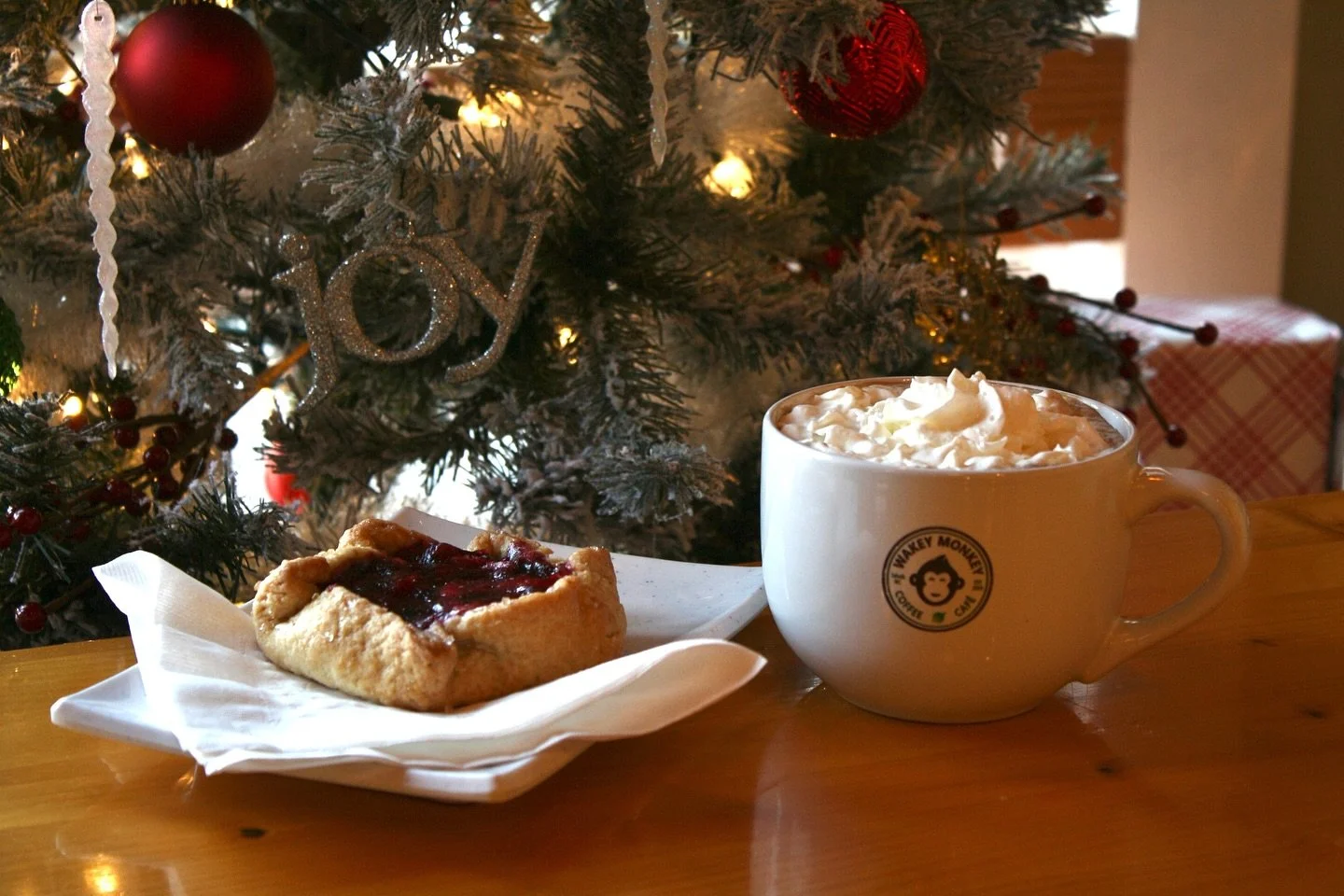 A plate with a cherry jam or pie square and a mug of hot chocolate topped with whipped cream, set on a wooden table in front of a decorated Christmas tree.