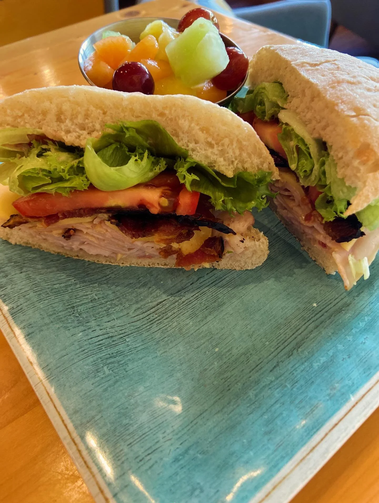 Close-up of a turkey and vegetable sandwich on a bun with lettuce, tomato, and bacon, served with a side of fresh fruit in a small bowl.